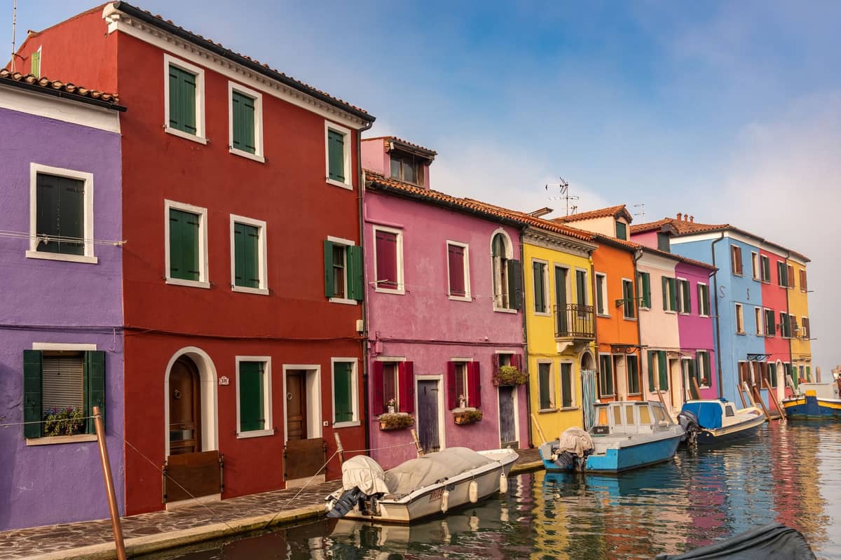Bunte Häuser entlang eines Kanals in Burano mit festgemachten Booten davor – ein Blick auf die ikonischen, leuchtenden Fassaden der venezianischen Laguneninsel.