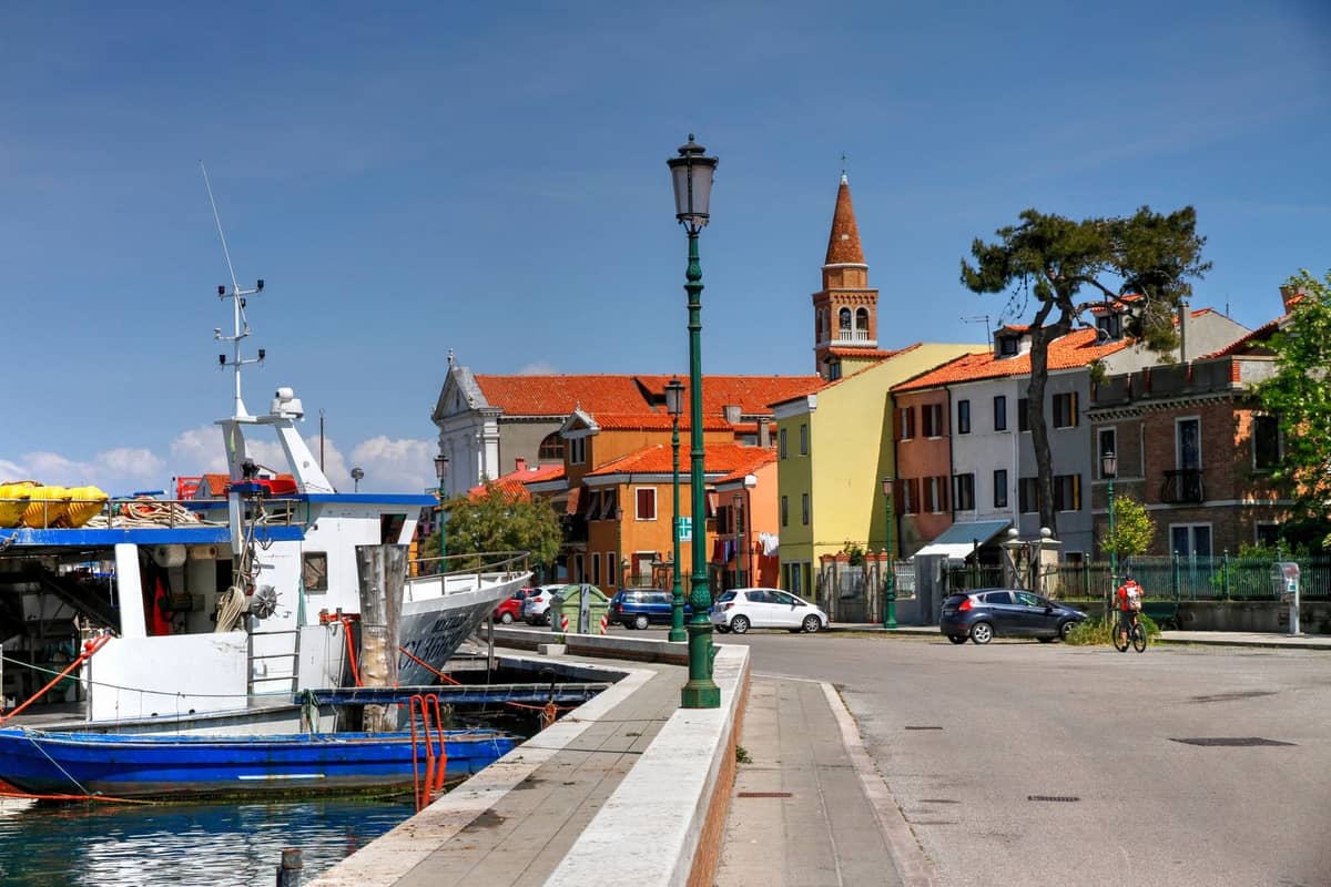 Fischerboot, das entlang einer Uferstraße mit bunten Häusern, Kirchturm und einigen Menschen in Pellestrina oder Chioggia, Lagune von Venedig, angelegt hat.