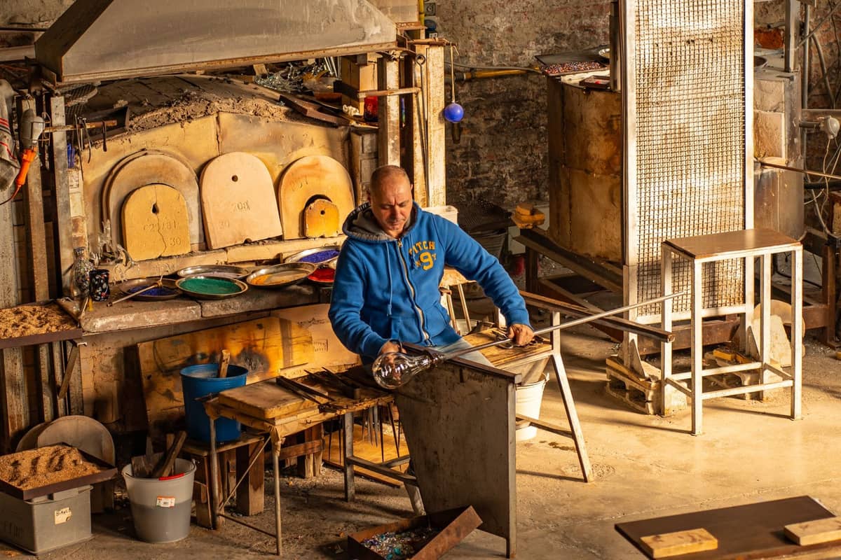 Glasmacher bei der Arbeit in einem traditionellen Murano-Ofenstudio, umgeben von Ausrüstung, Werkzeugen und einem glühenden Ofen.