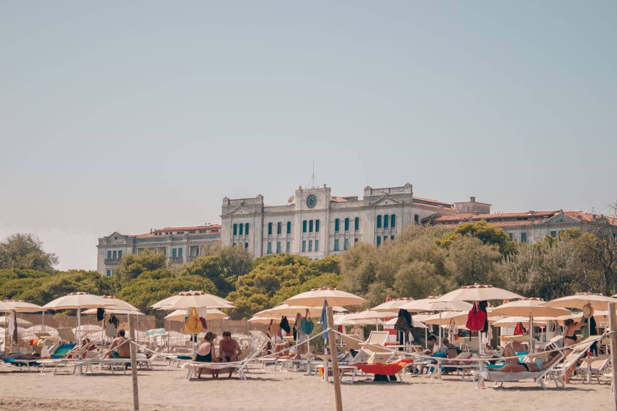 Menschen, die unter Sonnenschirmen an einem Sandstrand entspannen, mit einem prachtvollen Gebäude im Liberty-Stil und Bäumen im Hintergrund, am Lido di Venezia.