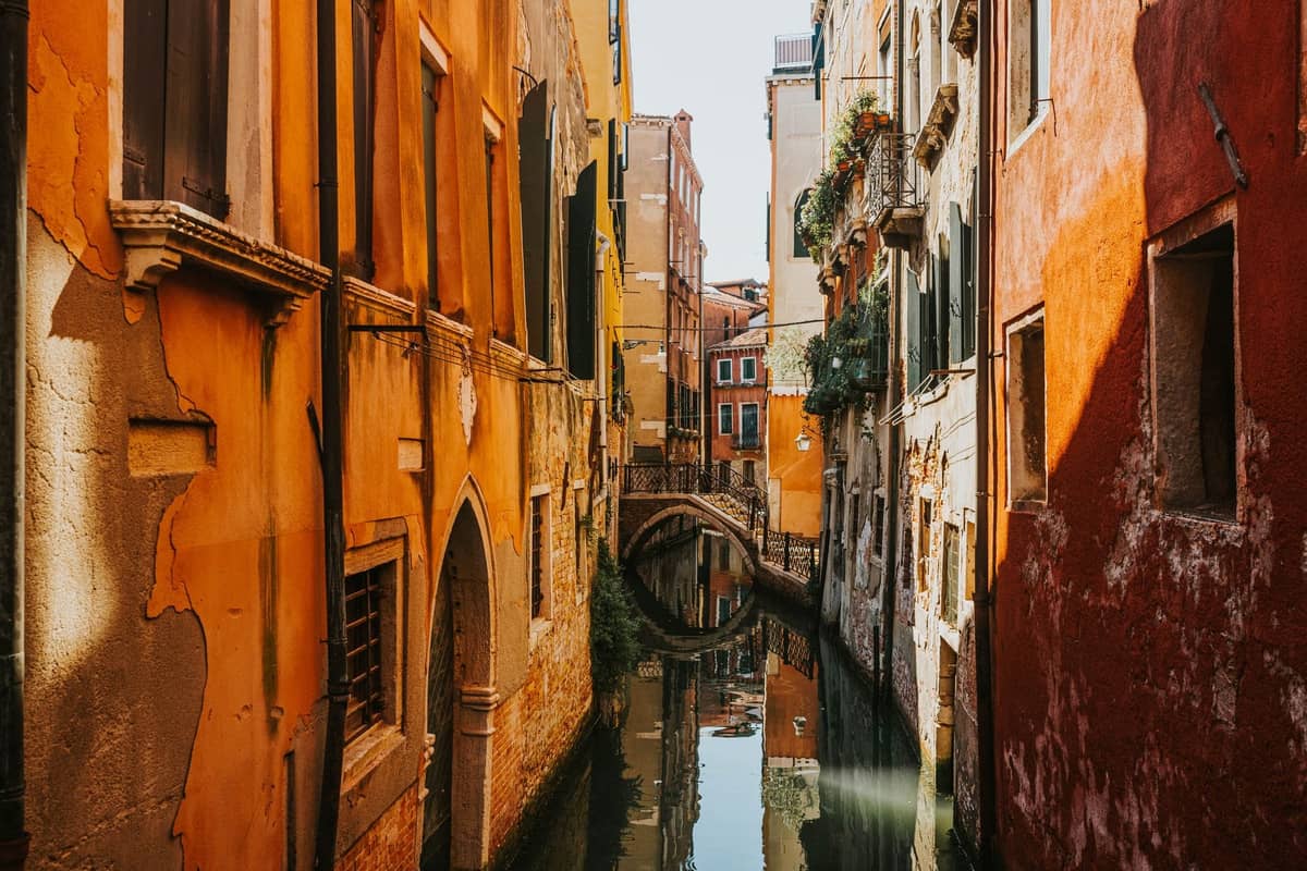 Schmaler Kanal in Venedig, gesäumt von bunten historischen Gebäuden und einer kleinen Fußgängerbrücke im Hintergrund.