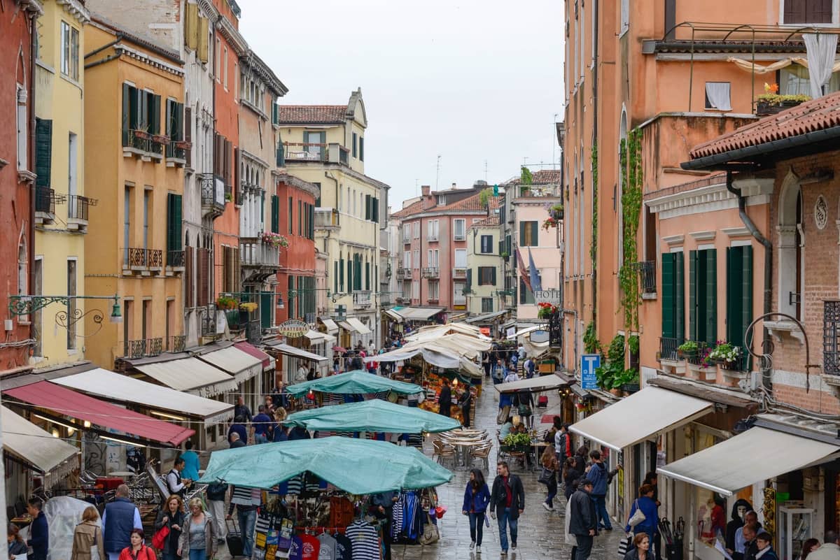 Belebte Einkaufsstraße in Venedig mit bunten Häusern, Marktständen und Passanten unter Markisen an einem bewölkten Tag.