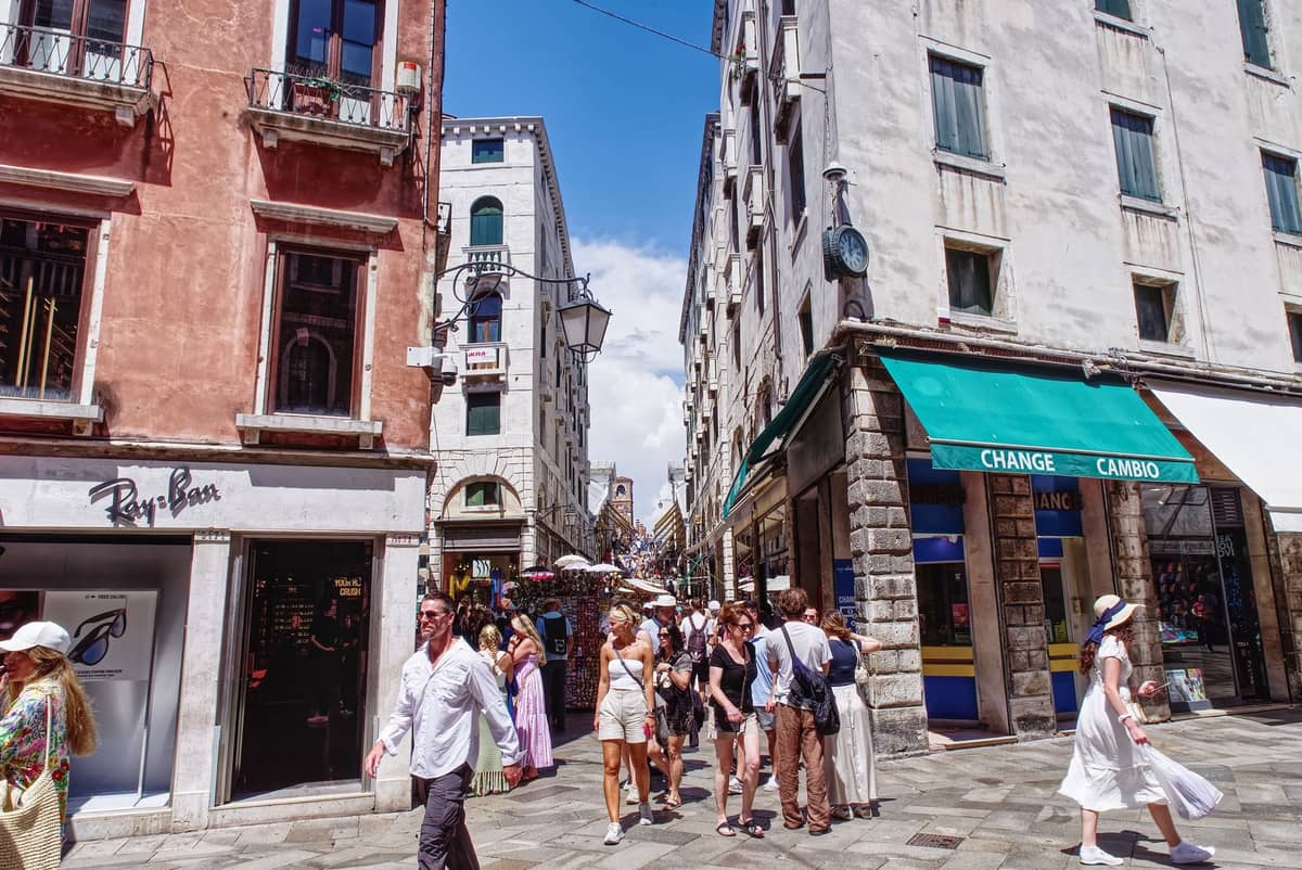 Belebte Straßenszene in Venedig mit Menschen, die an kleinen Geschäften vorbeigehen, keine Autos, umgeben von alten venezianischen Gebäuden und engen Gassen.