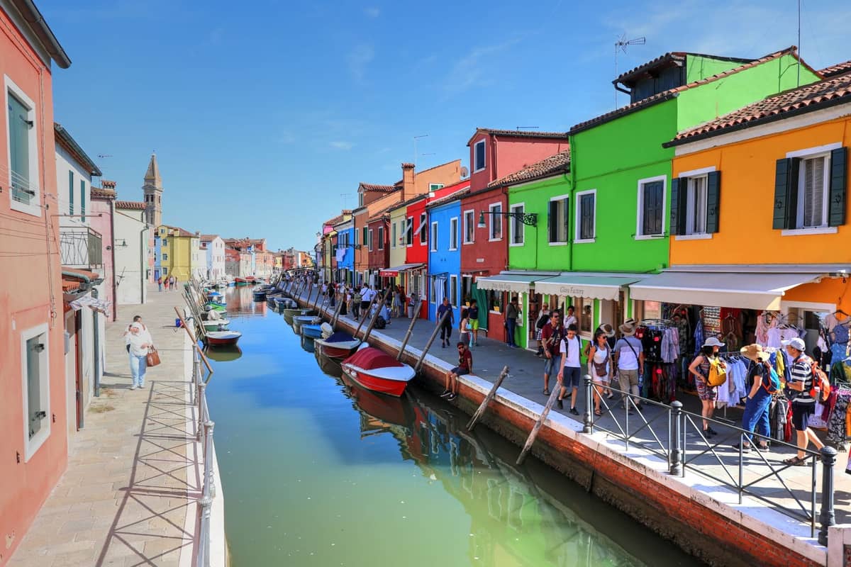 Bunte Gebäude säumen einen Kanal in Burano, mit Marktständen im Freien, Booten und Menschen, die am Wasser entlanggehen.