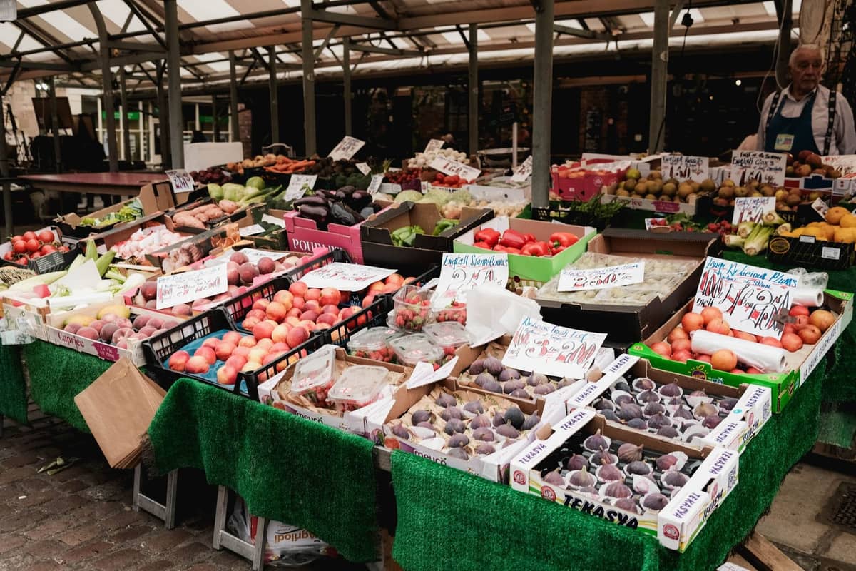 Frisches Obst und Gemüse auf einem venezianischen Marktstand mit handgeschriebenen Schildern und einigen Käufern im Hintergrund.