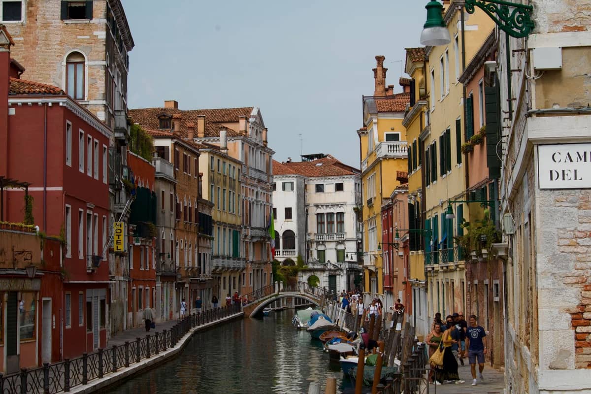 Venezianischer Kanal mit bunten Gebäuden, Steinweg, Menschengruppen und festgemachten Booten am Wasser unter einem bedeckten Himmel