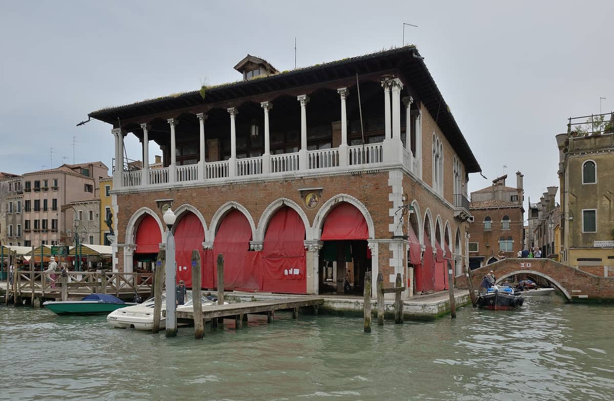 Blick auf das historische Fischmarktgebäude am Rialto in Venedig, mit roten Markisen, Bogenfenstern und am Kanal davor angedockten Booten.