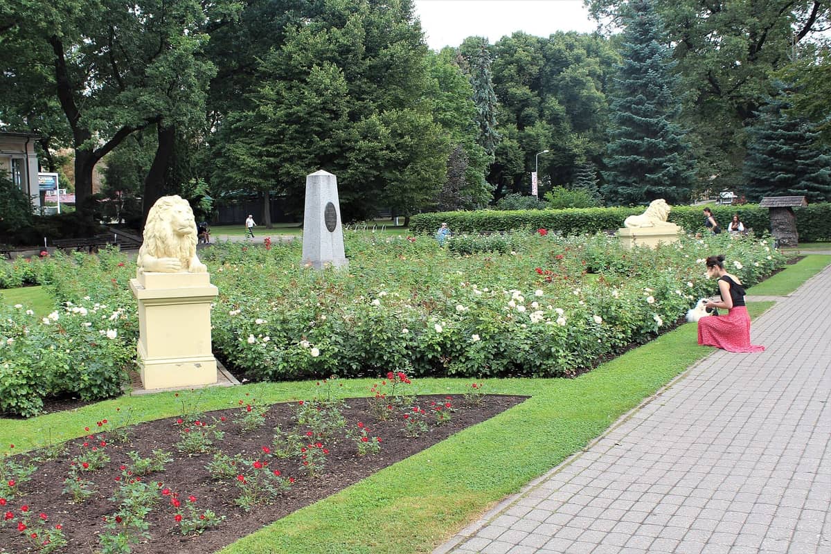 Weitläufiger Blick auf den Vērmane-Garten in Riga mit Blumenbeeten, Löwenstatuen, einem Denkmal, flanierenden und sitzenden Menschen sowie üppigen grünen Bäumen im Sommer.
