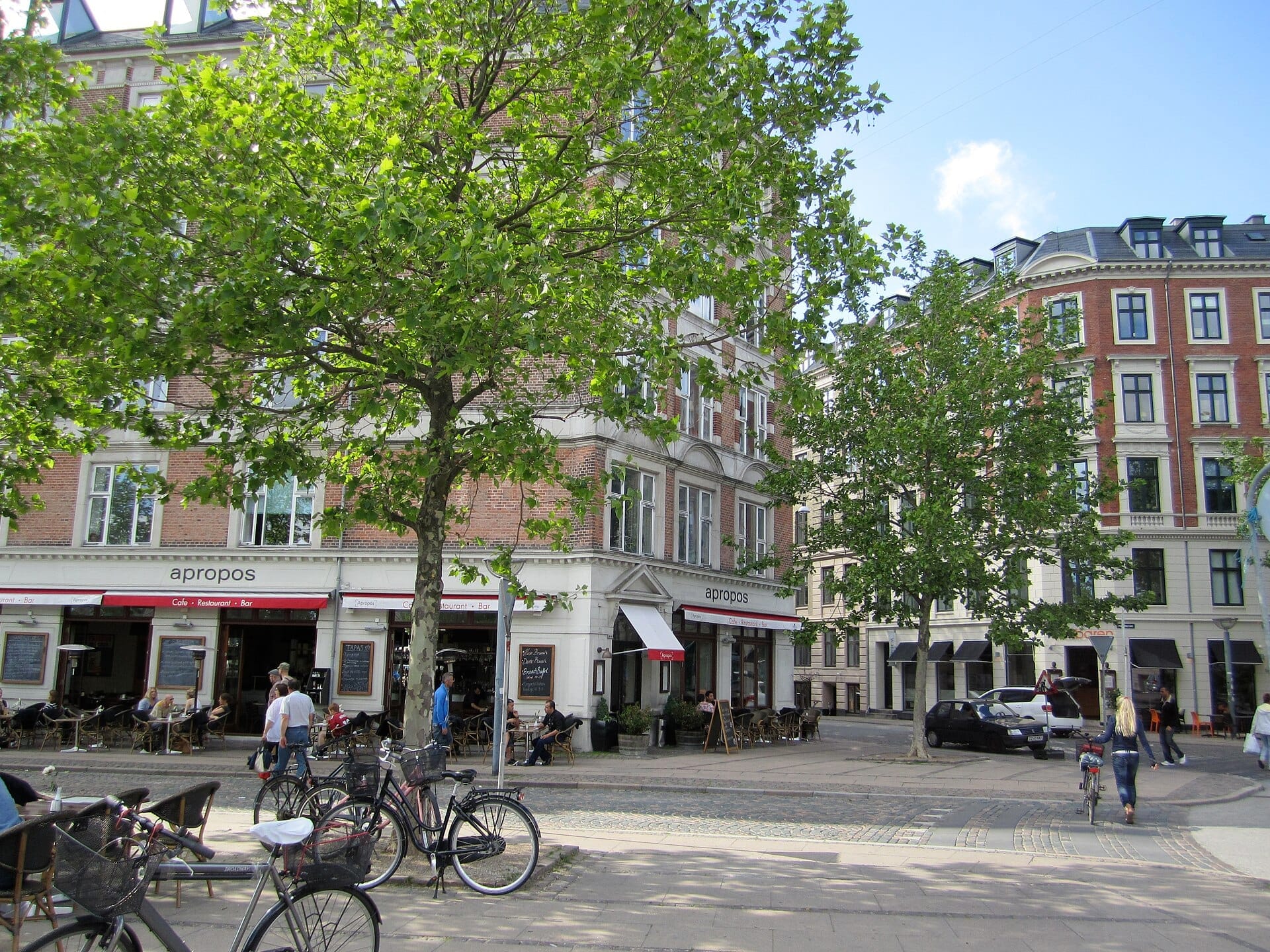 บรรยากาศคาเฟ่กลางแจ้งใน Vesterbro พร้อมผู้คนนั่งใต้ต้นไม้เขียวขจี จักรยานจอดอยู่ใกล้เคียง และอาคารอิฐประวัติศาสตร์เป็นฉากหลัง