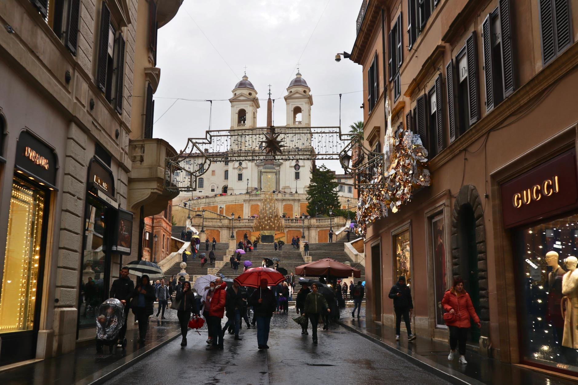People strolling along Via dei Condotti on a wet day, lined with luxury shops like Gucci and adorned with festive lights, with the Spanish Steps in the background.