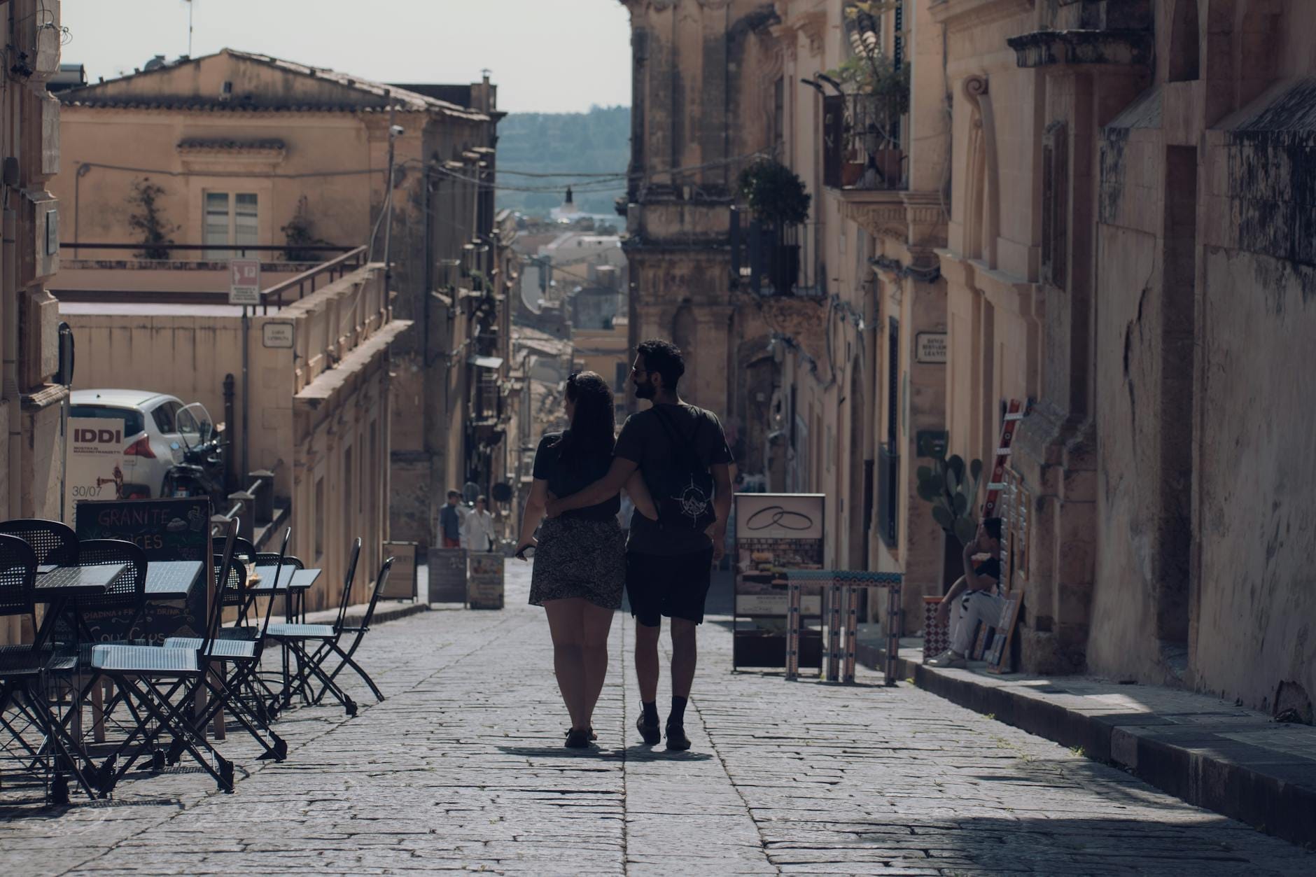 Ein Paar spaziert Arm in Arm durch eine sonnendurchflutete Barockstraße in Noto, gesäumt von eleganten historischen Gebäuden und Café-Tischen im Freien.
