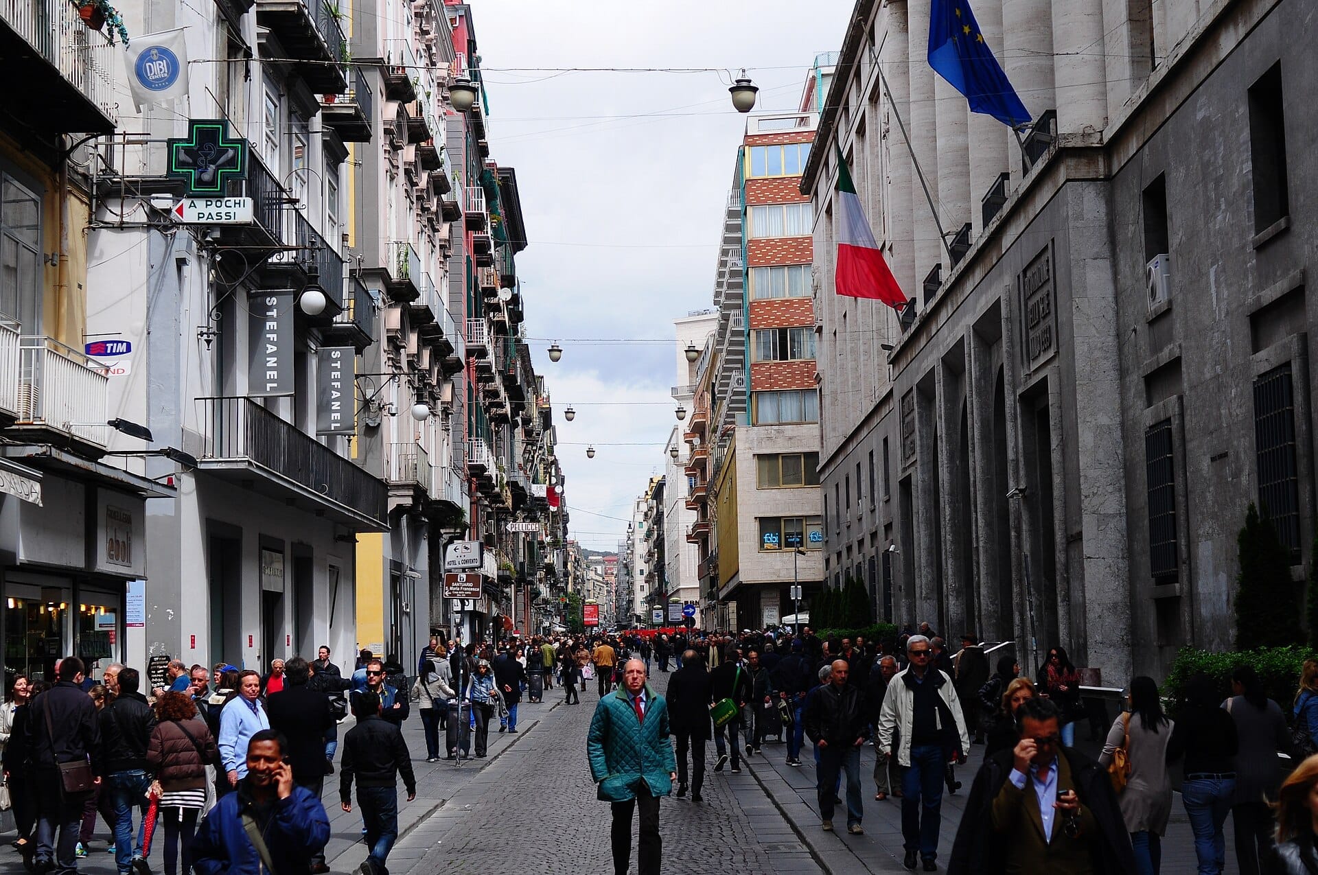 Una folla di persone passeggia su Via Toledo a Napoli, fiancheggiata da edifici storici, negozi e bandiere italiane su una vivace strada pedonale.
