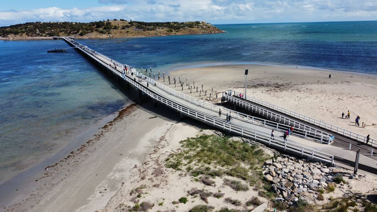 Ein langer Holzdamm führt unter hellem Himmel vom Sandstrand zur Granite Island, auf dem Menschen entlangspazieren – Victor Harbor.