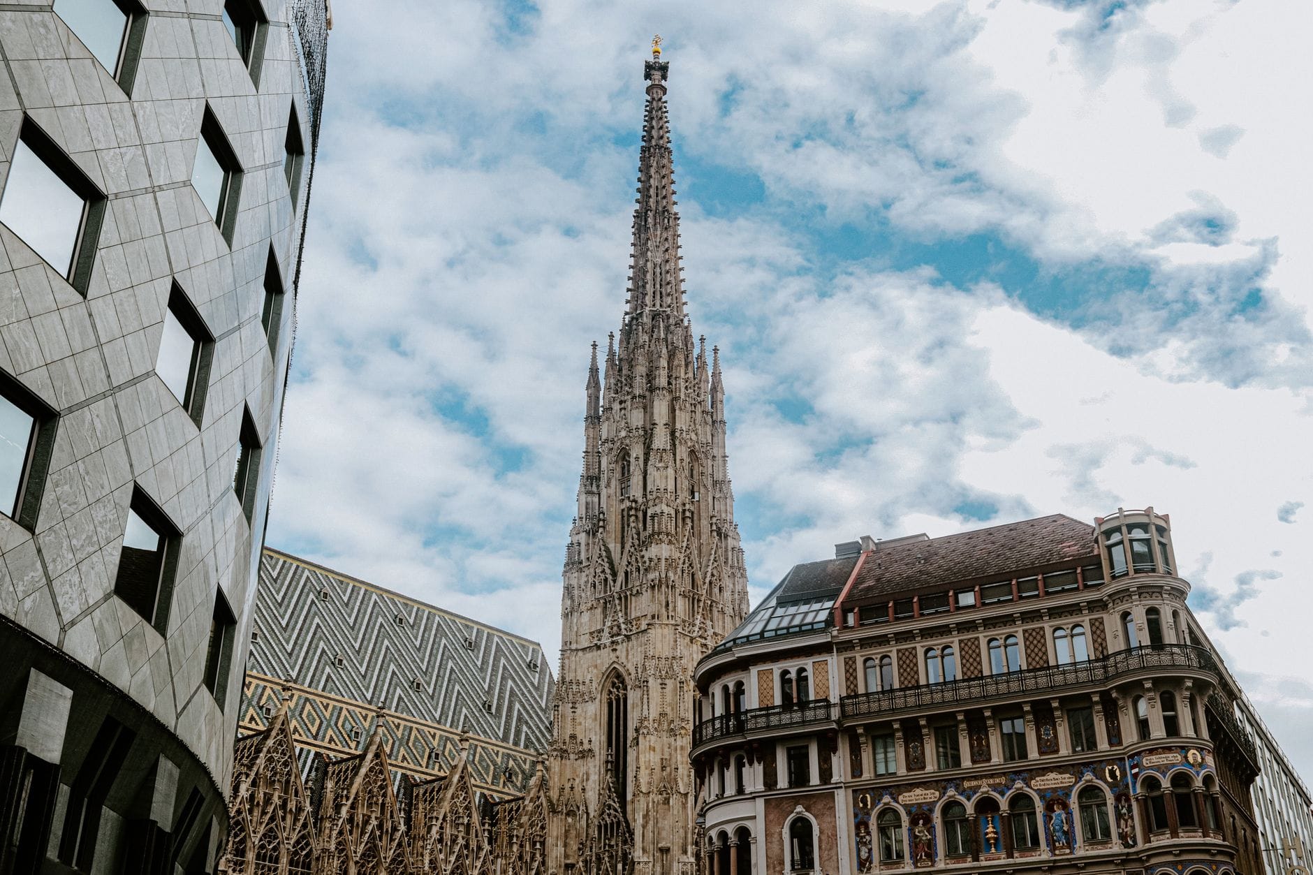 Vista a nivel de calle de la aguja y el techo de tejas coloridas de la Stephansdom en Viena, enmarcada por edificios históricos y modernos bajo un cielo luminoso.