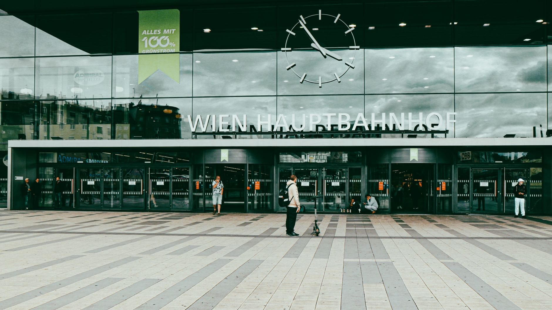 Haupteingang des Wien Hauptbahnhof mit Glasfassade, großer Uhr und Menschen vor dem Bahnhof