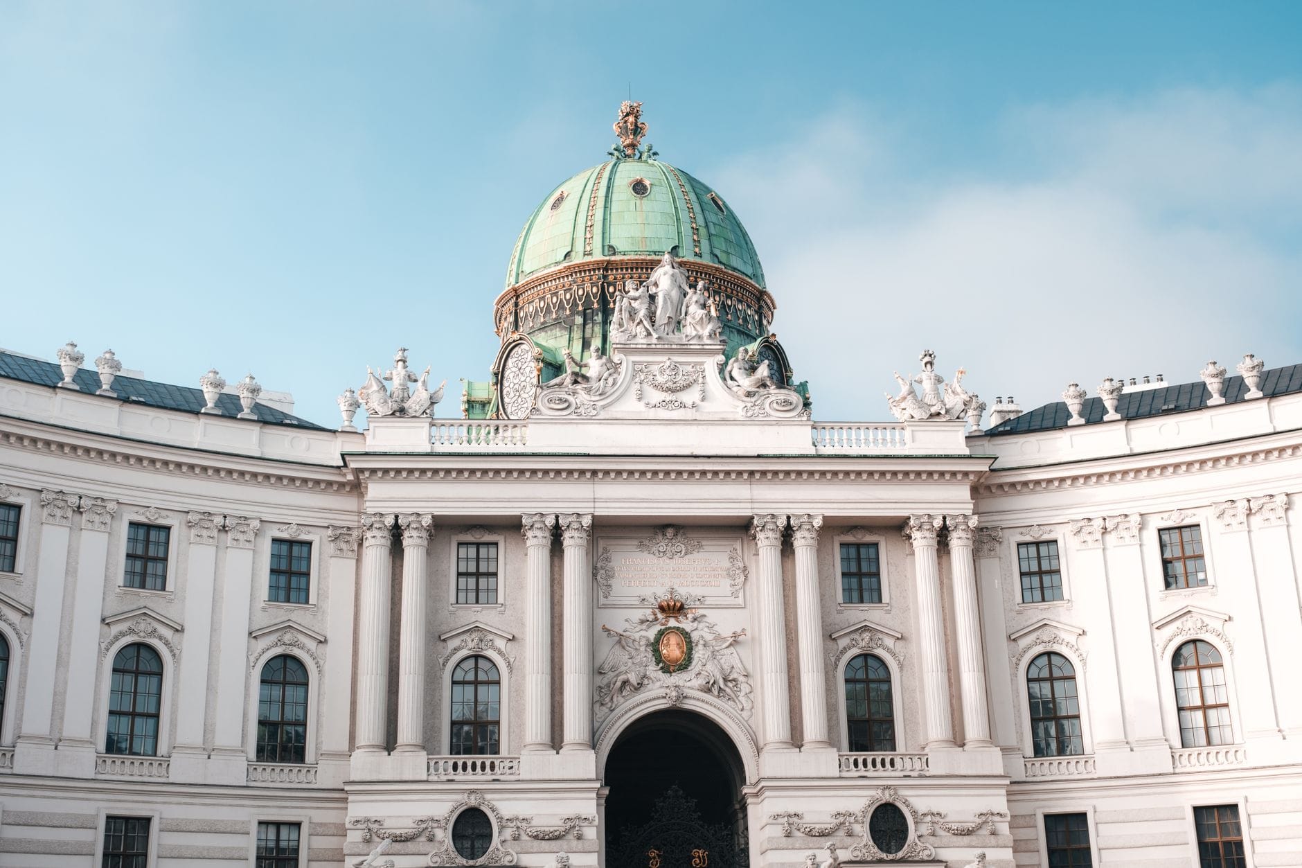 Vorderansicht des Hofburg-Palastes in Wien mit seiner verzierten grünen Barockkuppel und der detaillierten weißen Steinfassade vor blauem Himmel.