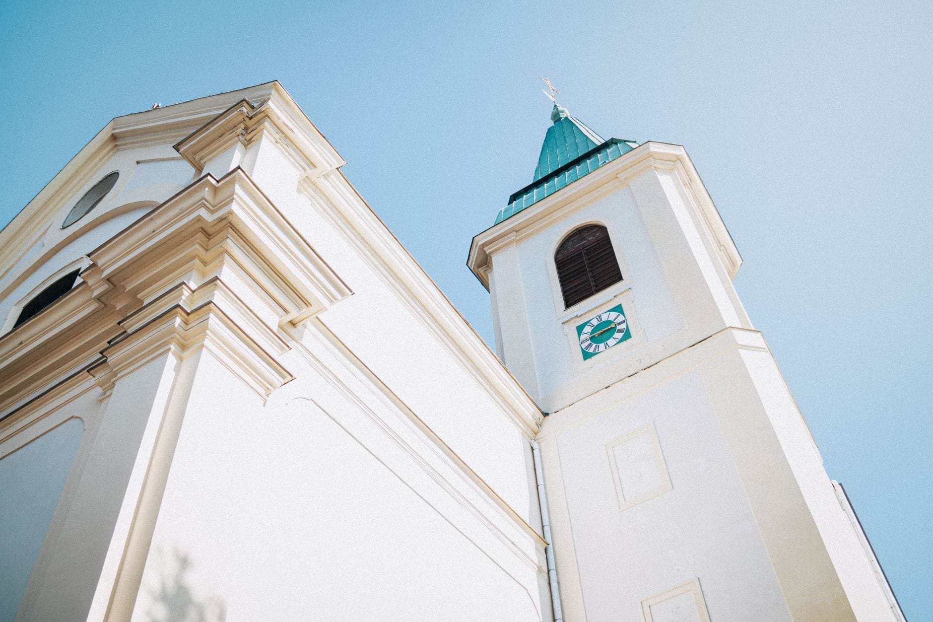 Blick nach oben auf eine schlichte weiße Kirche mit grünem Kupferdach und Uhrturm vor blauem Himmel.