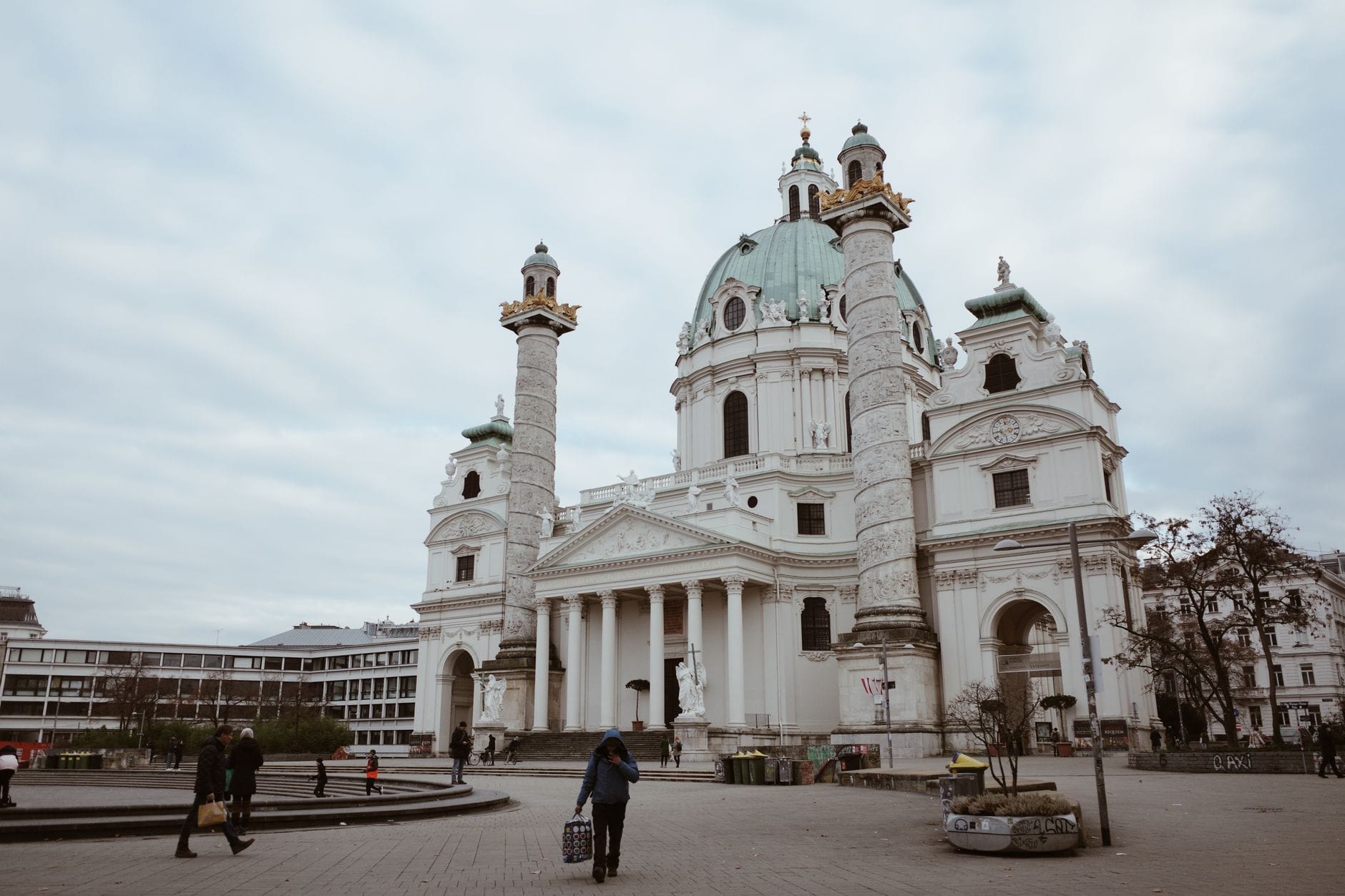 Weiter Blick auf die Wiener Karlskirche, eine prächtige Barockkirche mit grüner Kuppel und aufwendigen Säulen auf einem Stadtplatz unter bewölktem Himmel.
