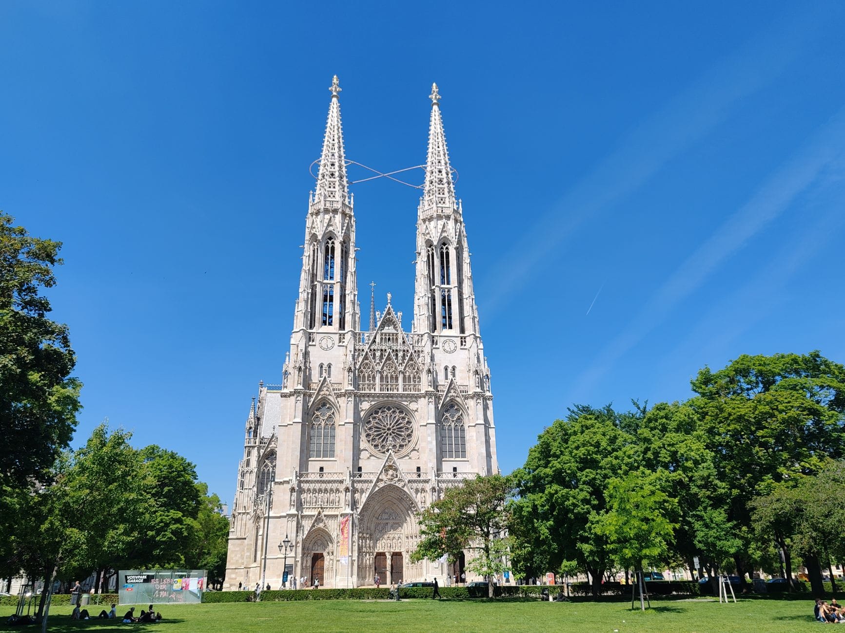 Weitwinkelansicht der Votivkirche mit ihren zwei hohen neugotischen Türmen, großem Rosettenfenster und grünem Park davor, unter einem klaren blauen Himmel.