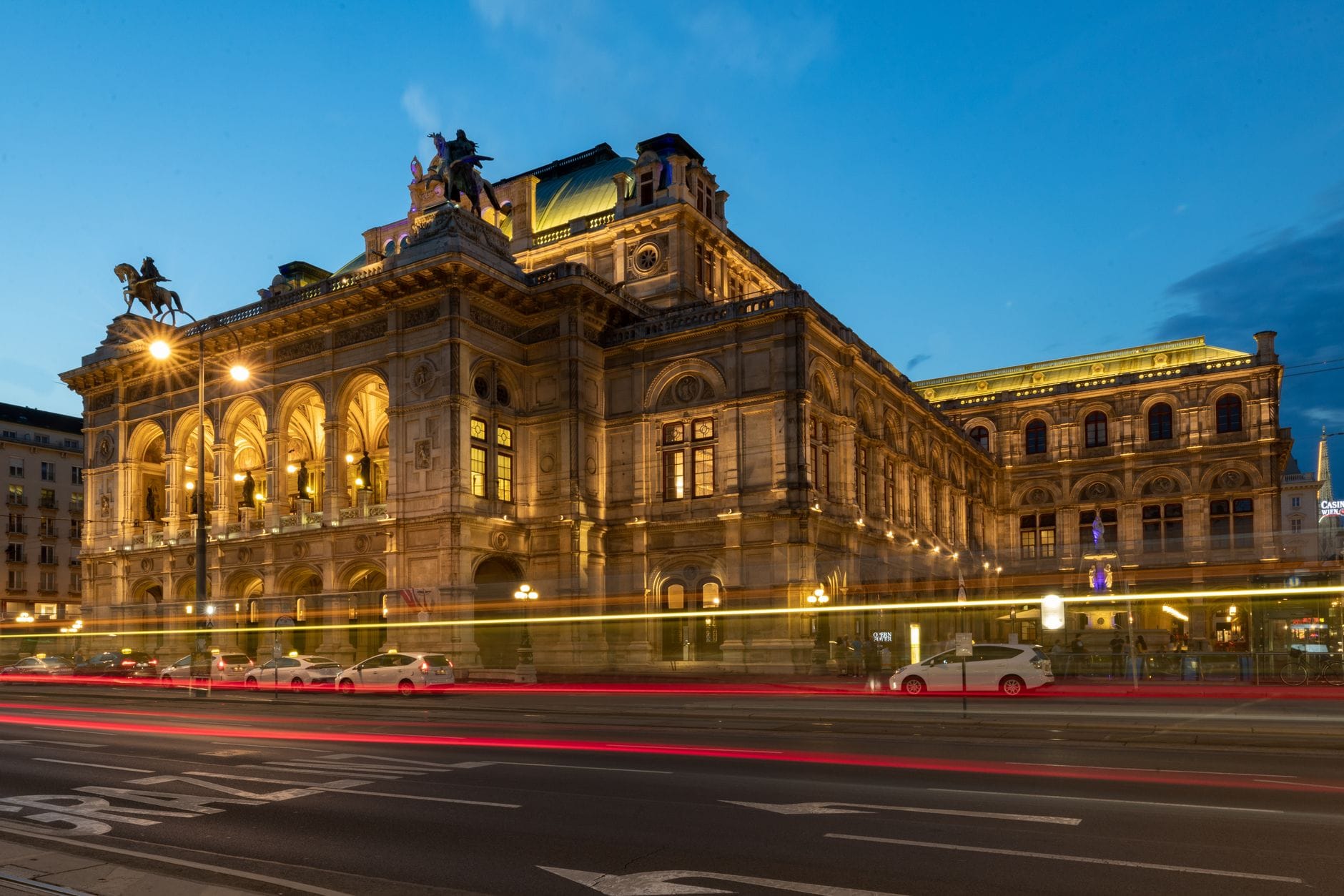 Weitwinkelaufnahme der Wiener Staatsoper in der Abenddämmerung, prachtvoll beleuchtet mit Lichtstreifen vorbeifahrender Autos davor – eine magische Stadtkulisse.