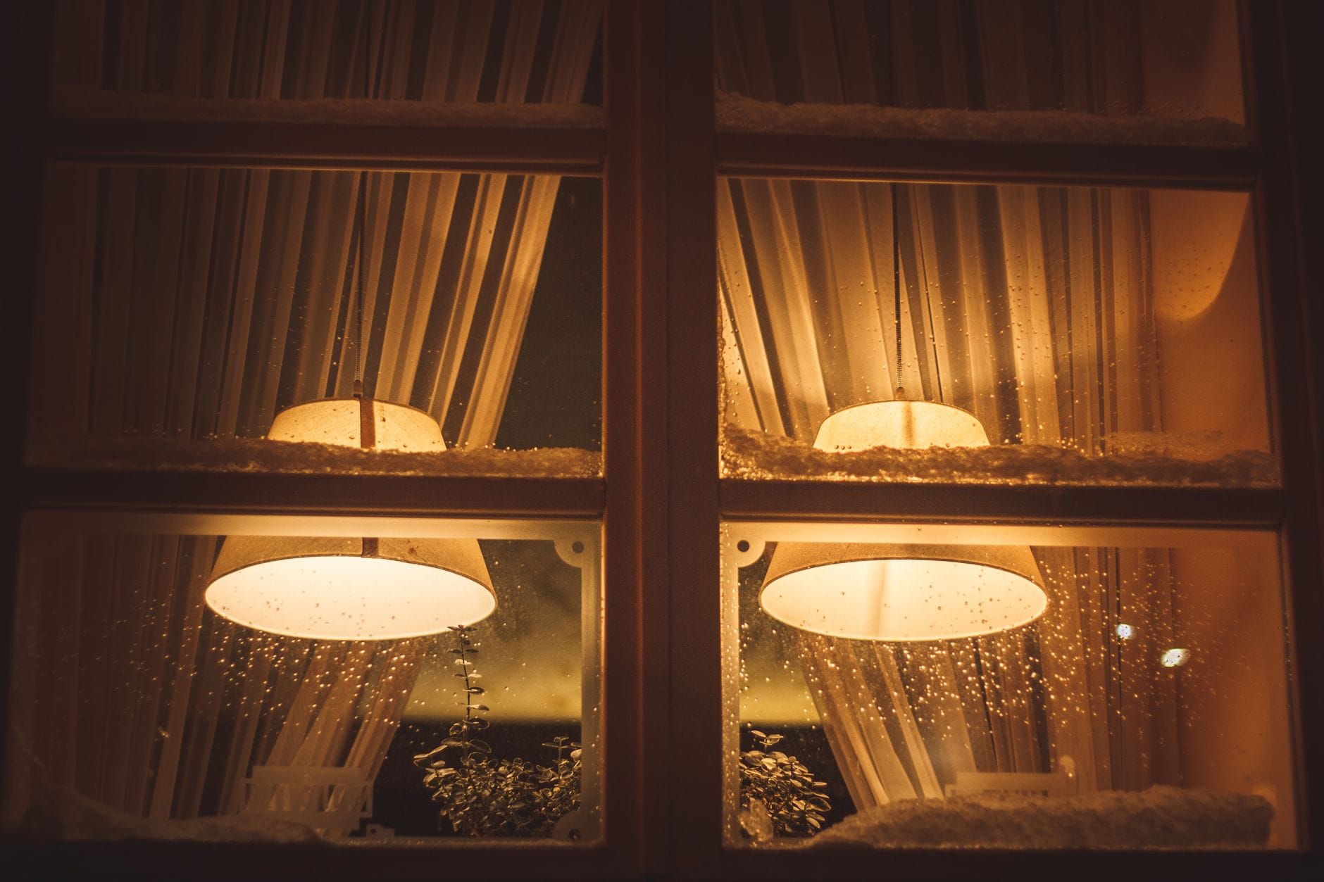 Una vetrina di caffè illuminata calorosamente con lampade, neve leggera e gocce di pioggia sul vetro, che evoca un'accogliente atmosfera invernale all'interno.