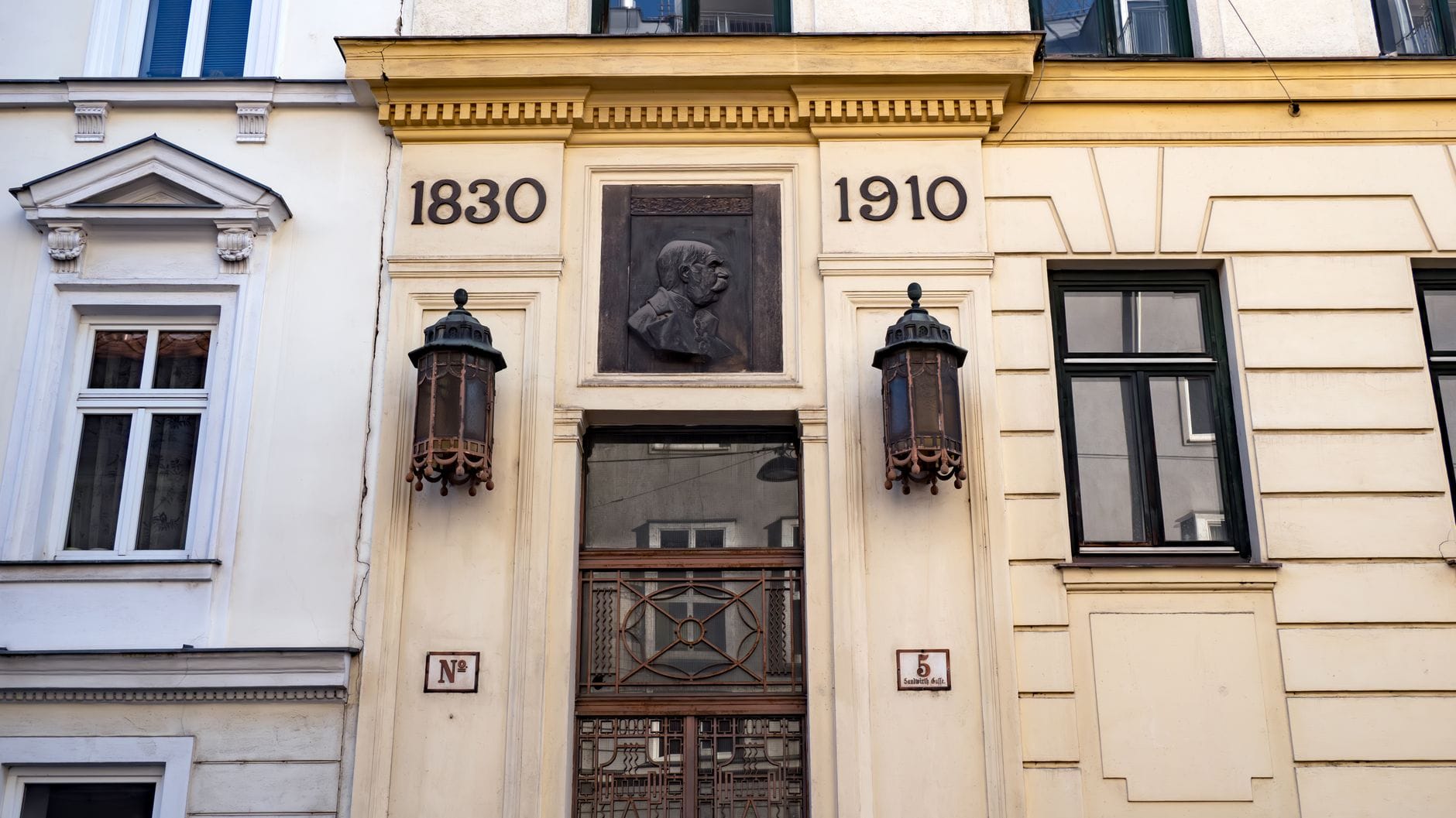 Elegante Fassade eines Wiener Gebäudes mit historischen Laternen, einem bronzenen Portraitrelief und den Jahreszahlen 1830 und 1910 über dem Eingang.