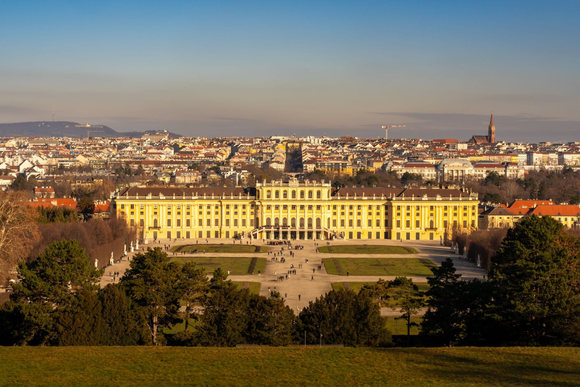 Panoramablick auf Schloss Schönbrunn in Wien mit dem formalen Garten, der Stadtsilhouette im Hintergrund und strahlend blauem Himmel.