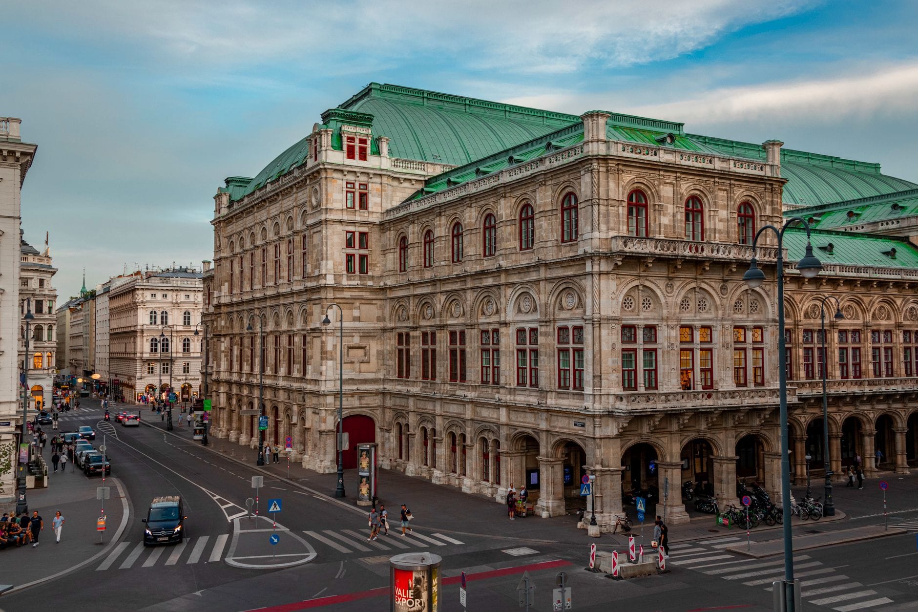 Vista ampia dell'Opera di Stato di Vienna sulla Ringstrasse con traffico, pedoni e grandi edifici storici sotto un cielo azzurro.