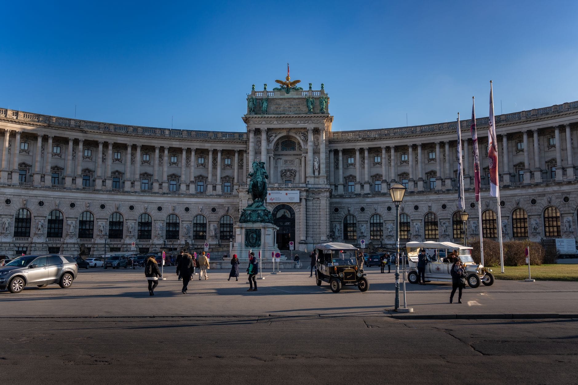 Vista ampia del Palazzo Hofburg a Vienna con statua equestre davanti, turisti e carrozze in primo piano, sotto un cielo azzurro limpido.