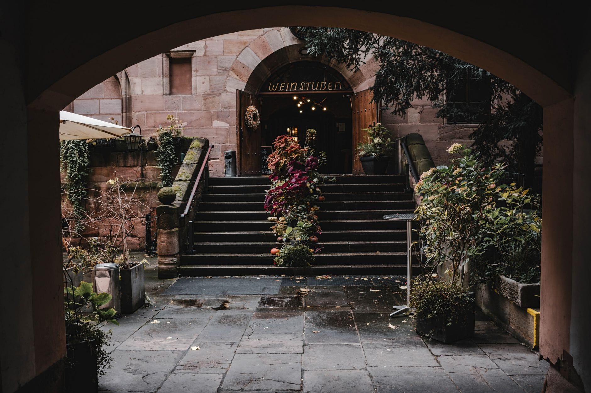 Arched stone entrance to a traditional wine tavern surrounded by lush plants, with steps leading up to a cozy courtyard setting.