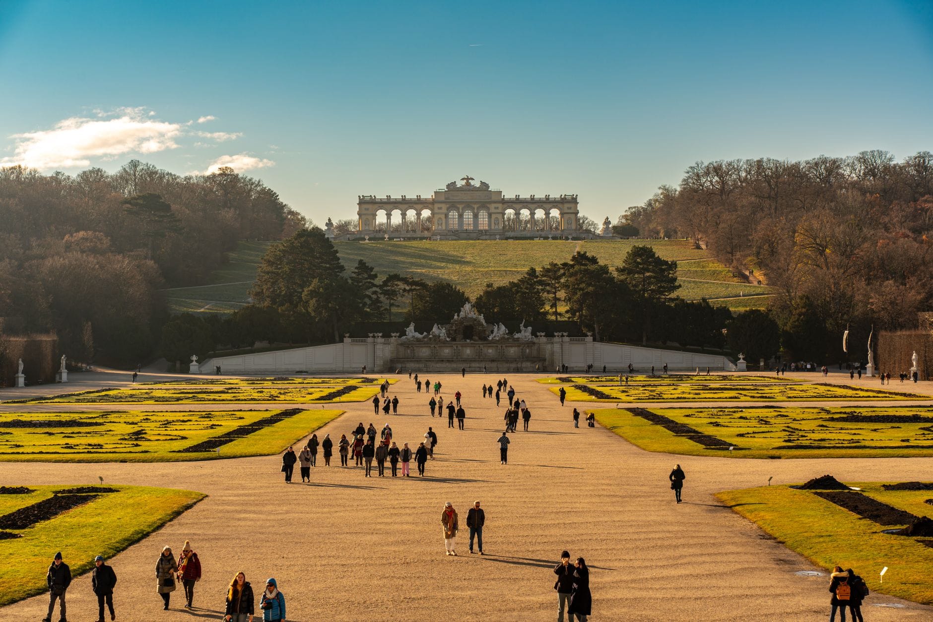 Menschen spazieren auf breiten Kieswegen durch Stadtgärten, im Hintergrund die bewaldeten Hügel des Wienerwalds und das historische Gloriette-Gebäude unter klarem Himmel.