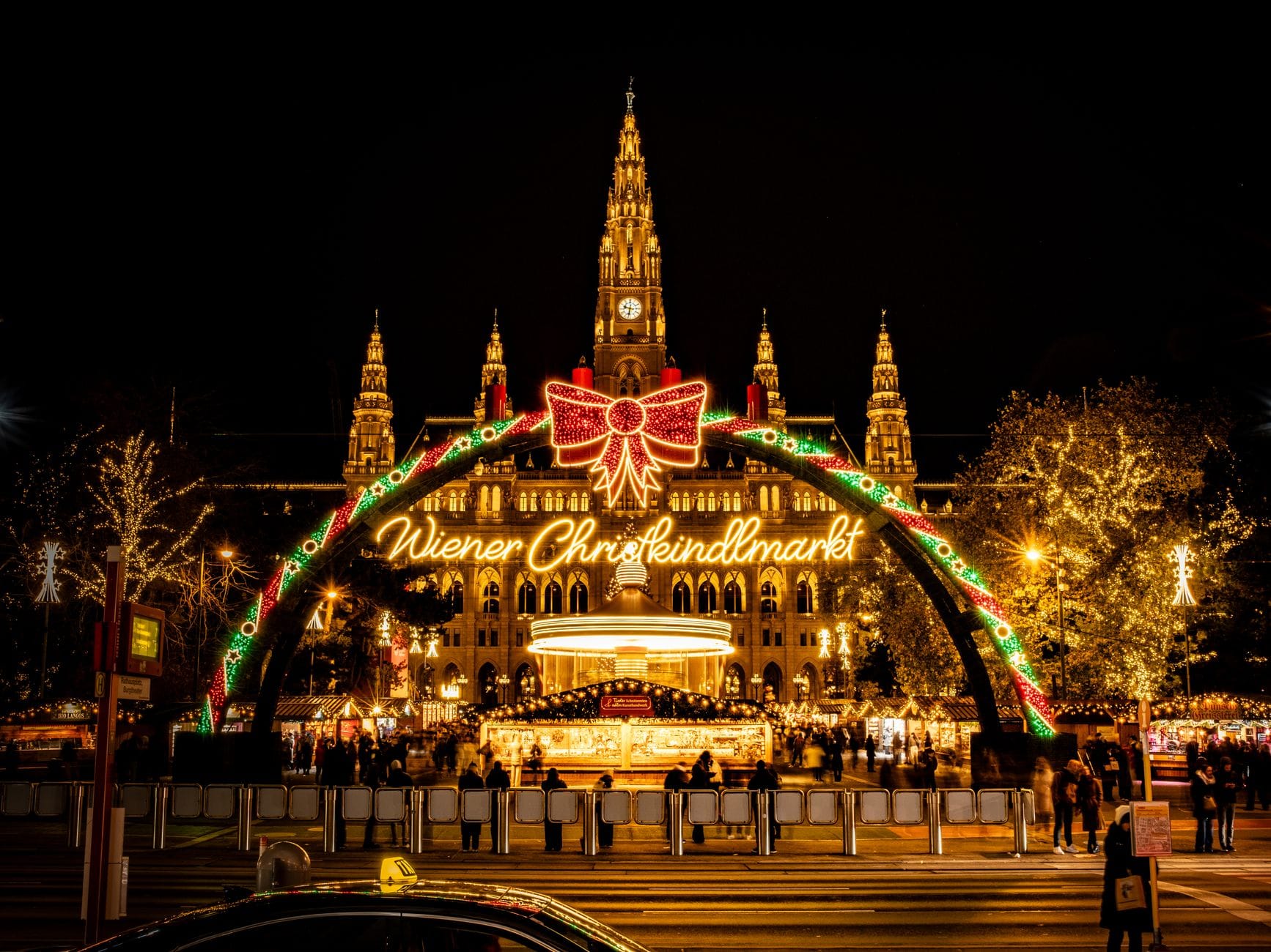 Vista notturna del Municipio di Vienna con l'insegna illuminata dell'ingresso del Wiener Christkindlmarkt e i vivaci stand del mercatino di Natale.