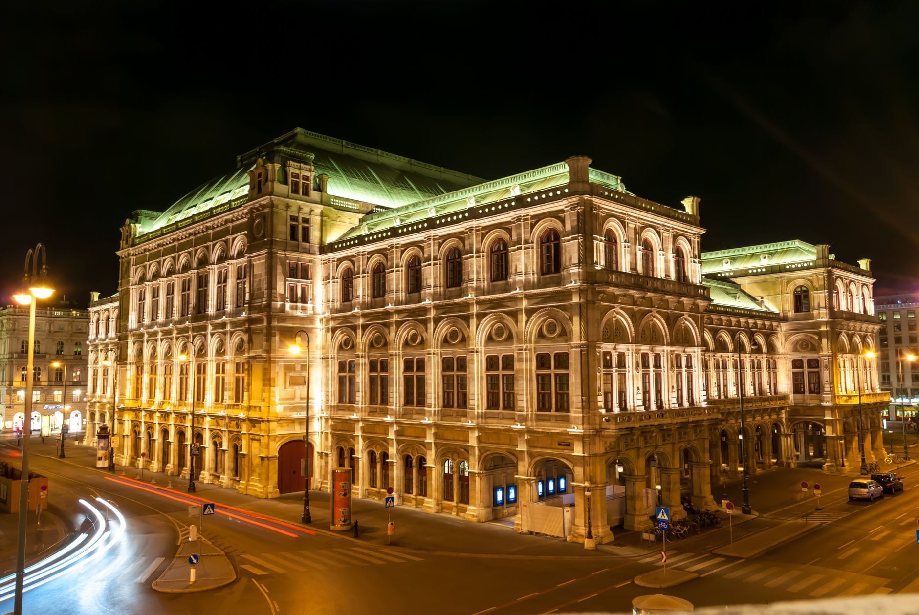 Edificio iluminado de la Ópera Estatal de Viena por la noche, con rastros de luz del tráfico en calles vacías de la ciudad.