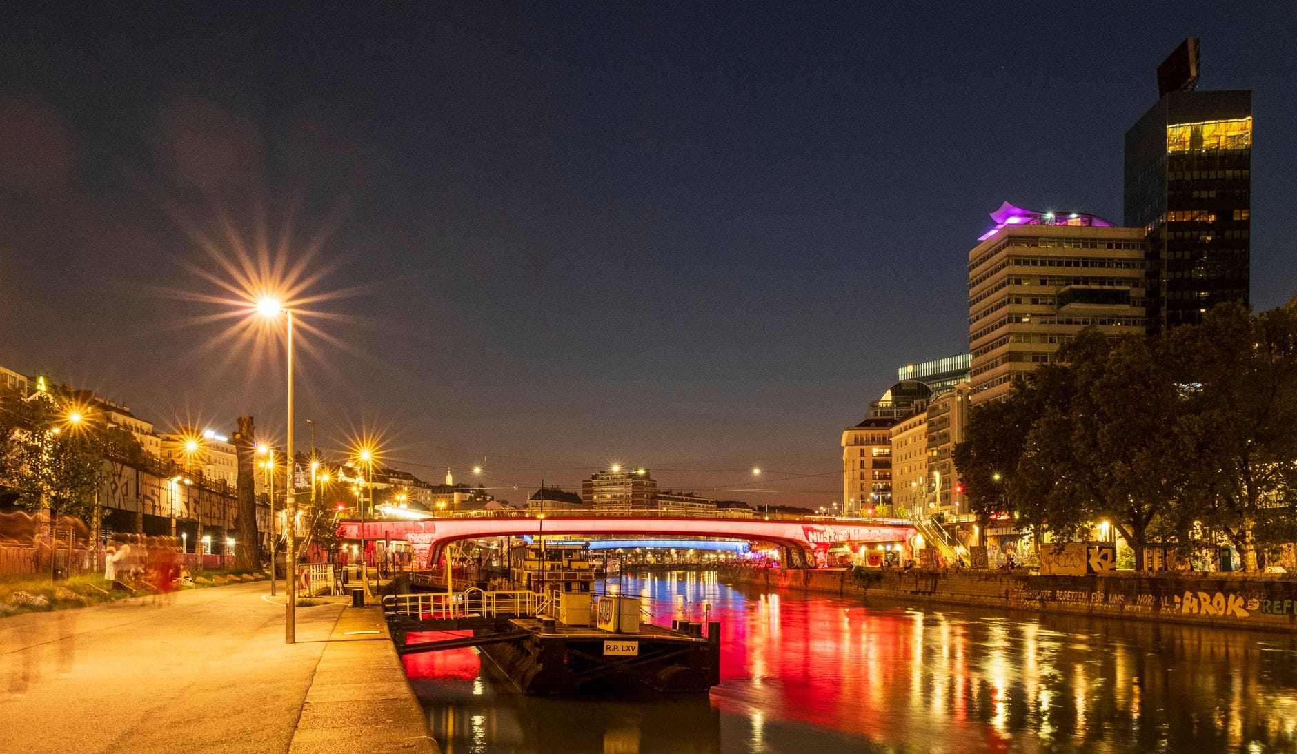 Vista nocturna de Viena a lo largo del Canal del Danubio con edificios iluminados y reflejos en el agua.