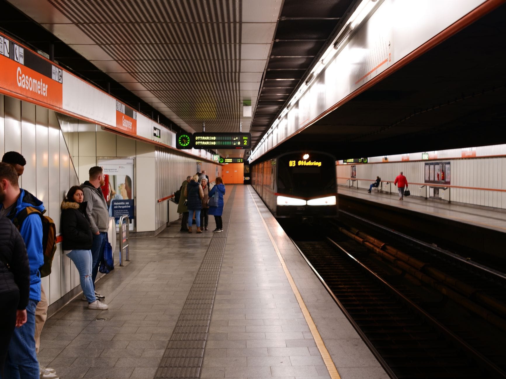 Personas esperan un tren de metro en la estación de U-Bahn Gasometer de Viena, con un tren llegando en la vía opuesta.