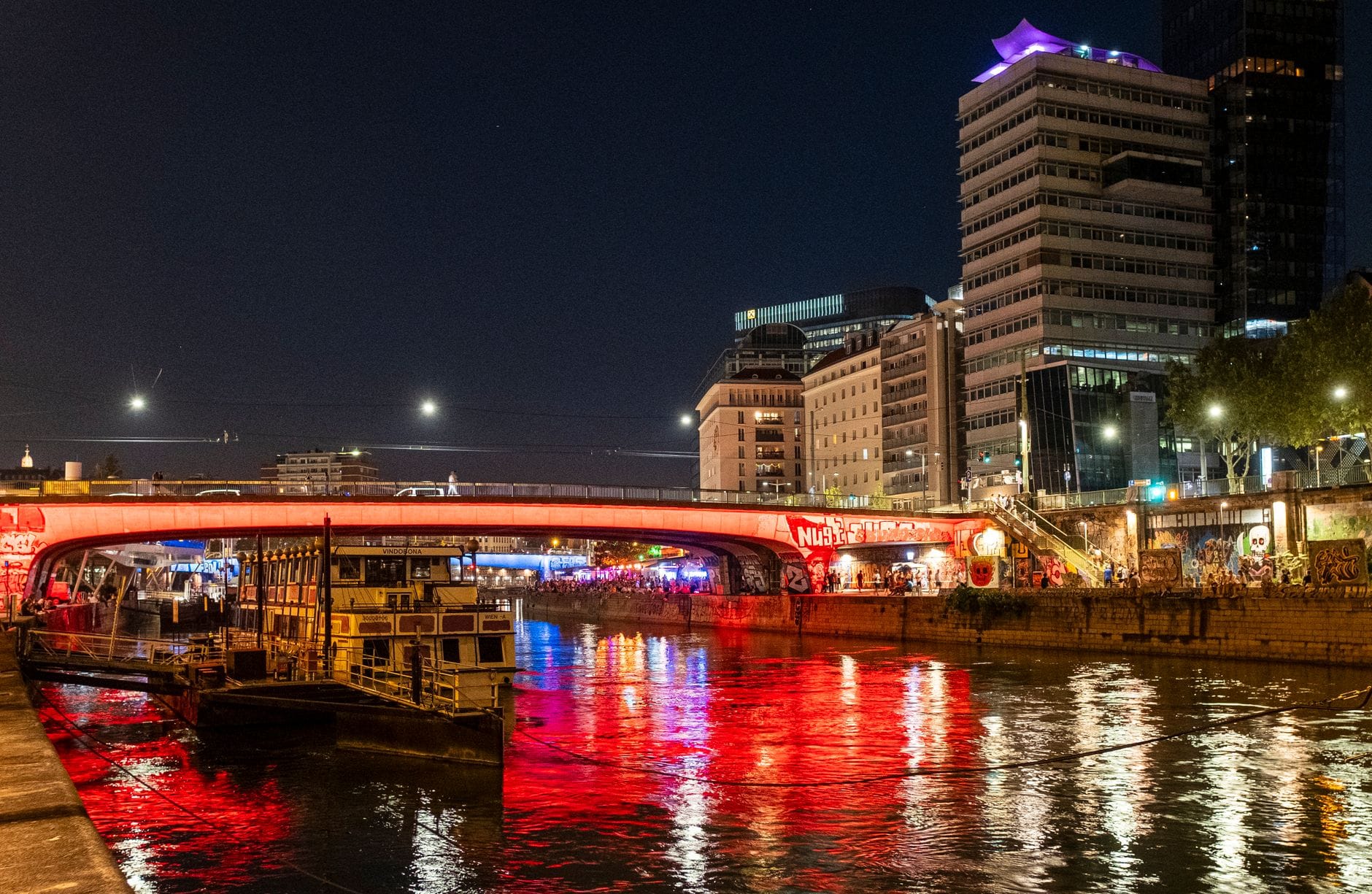 El canal del Danubio de Viena de noche, iluminado con luces de colores y reflejos, personas reunidas a lo largo del malecón para el ocio nocturno de verano.