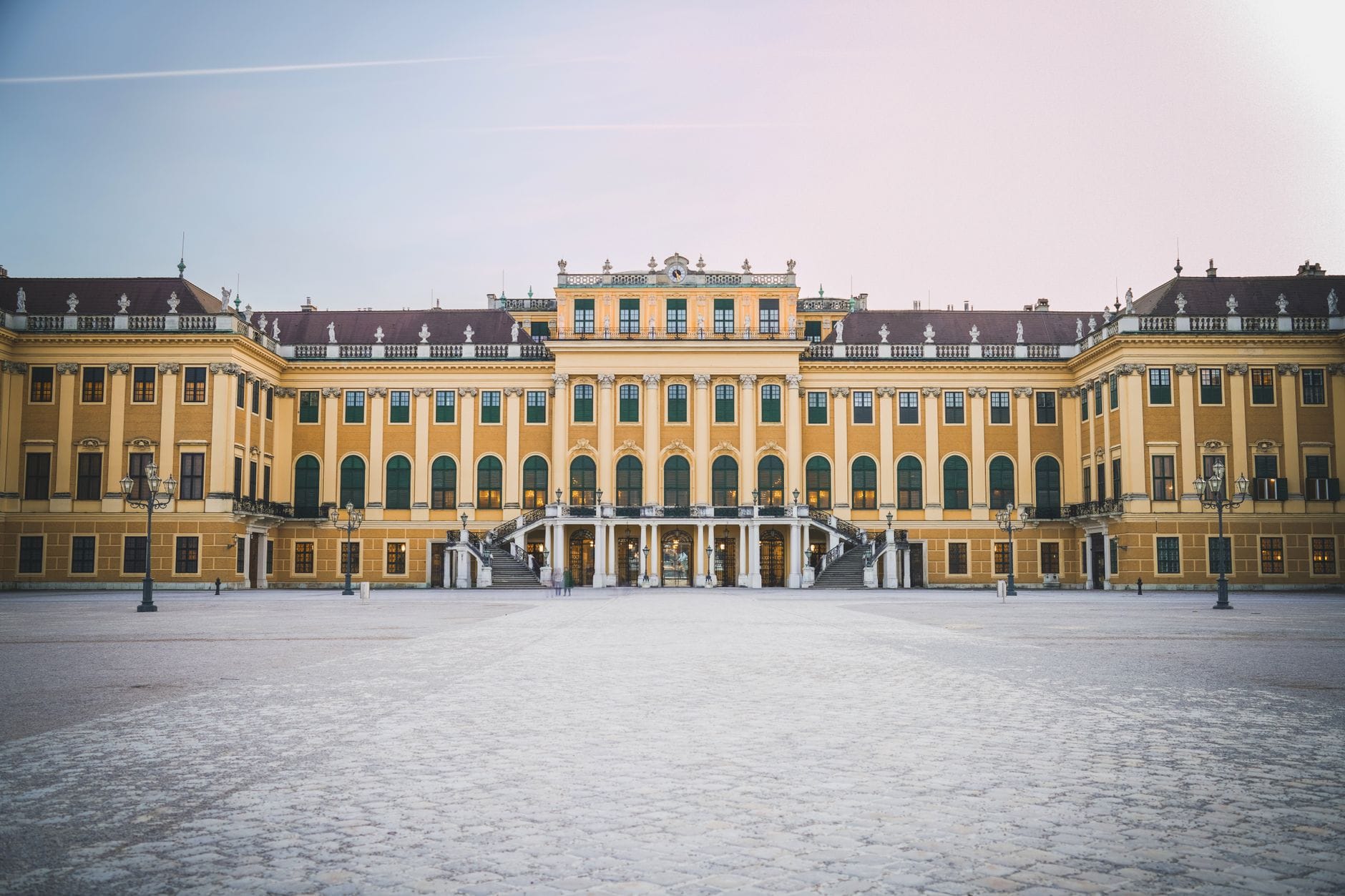 Die prachtvolle gelbe Fassade des Schlosses Schönbrunn in Wien, weitwinklig fotografiert unter einem sanften blau-rosa Himmel mit leerem Vorplatz im Vordergrund.