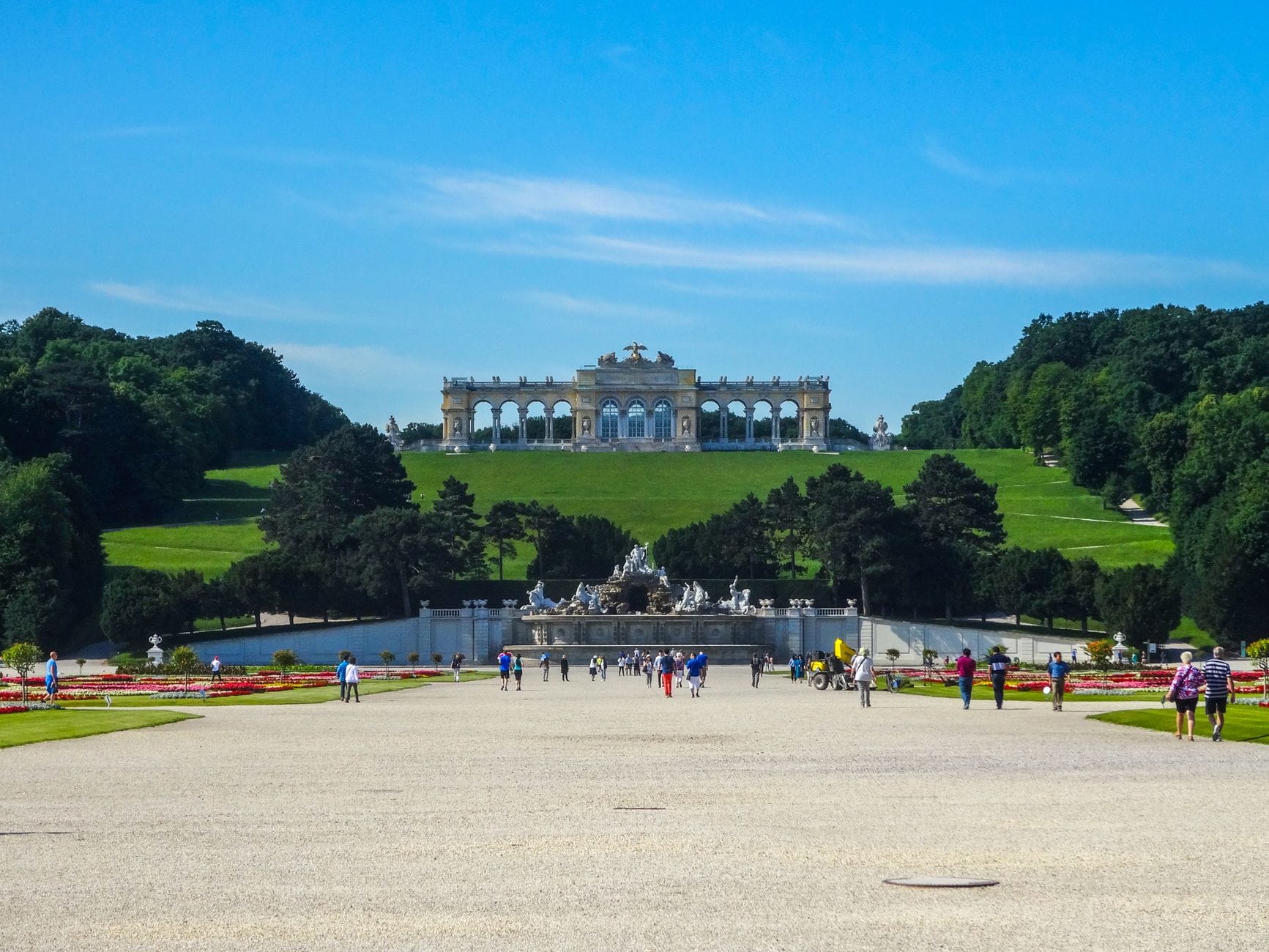Blick entlang der Hauptallee des Schönbrunner Gartens mit dem Gloriette-Pavillon auf dem Hügel, grünen Rasenflächen, Blumenbeeten und Spaziergängern.