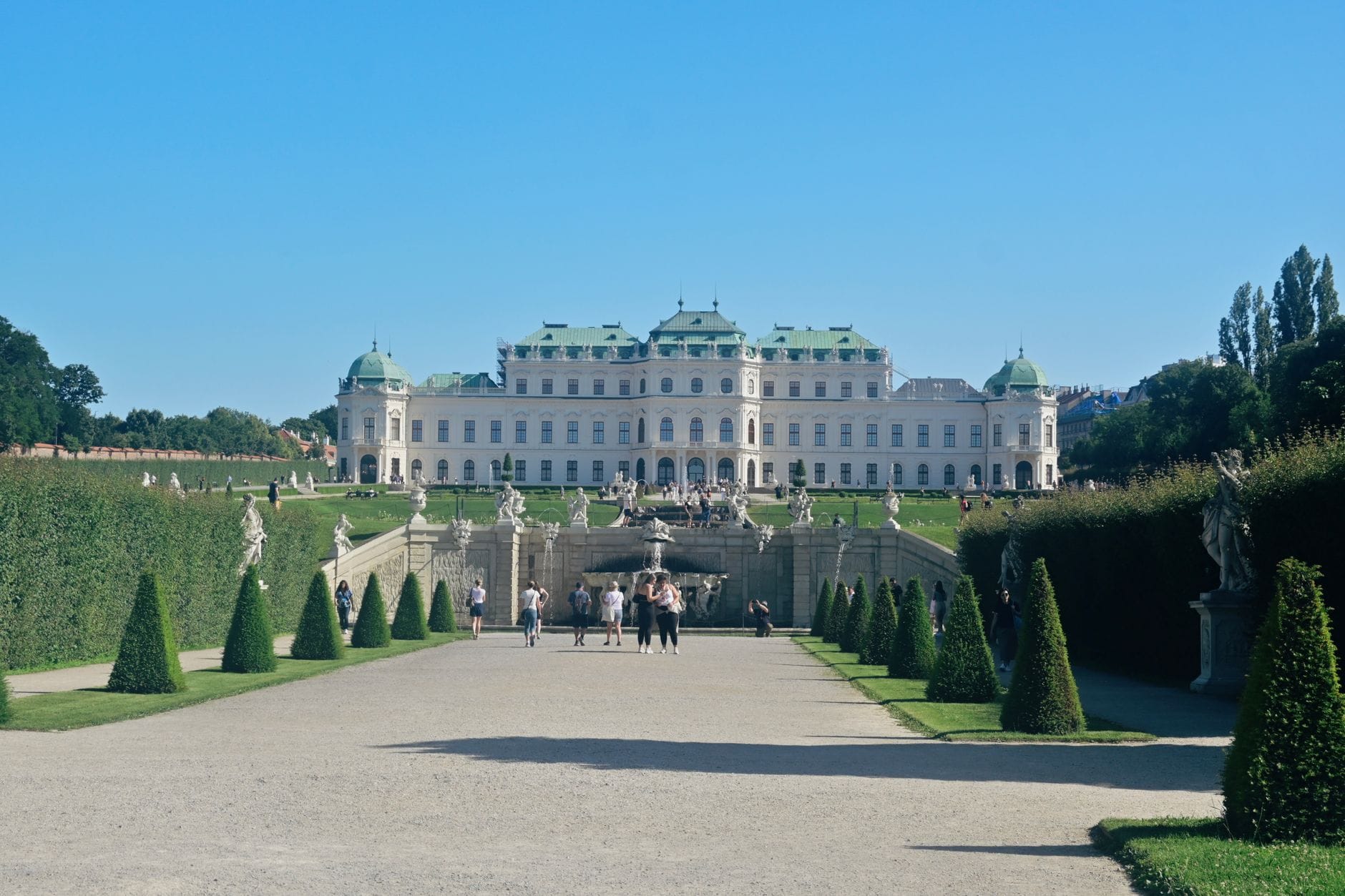 Frontale Ansicht des Oberen Belvedere-Palastes in Wien mit formellen Gärten, Statuen und sorgfältig gestutzten Hecken unter einem klaren blauen Himmel.