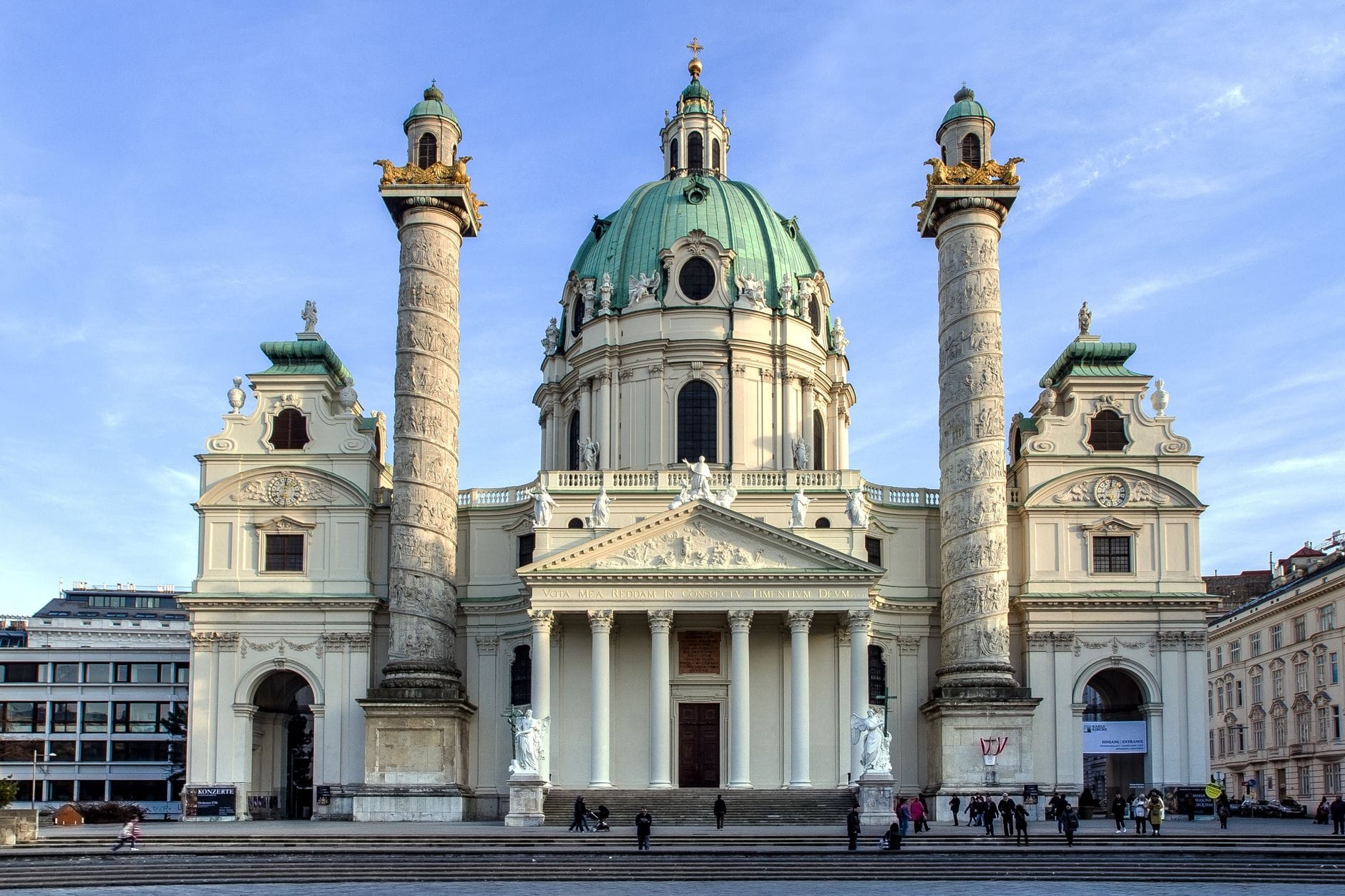 Weitwinkelansicht der Wiener Karlskirche mit ihrer barocken Kuppel und den zwei Säulen an einem klaren Tag, mit Menschen davor.