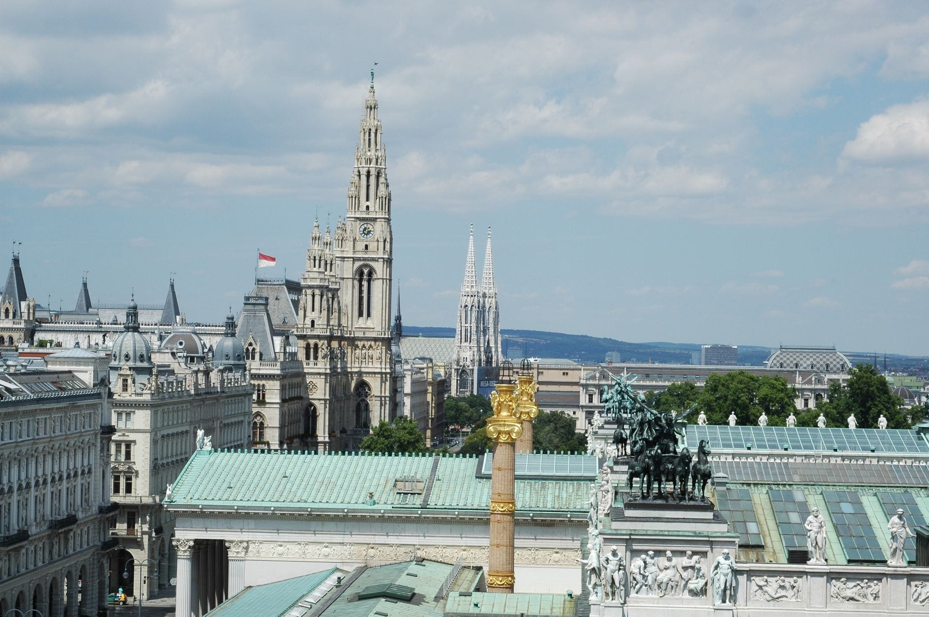 Weiter Blick über die Wiener Skyline mit den markanten Türmen des Rathauses und der Votivkirche, vor blauem Himmel und umgeben von historischer Architektur und sommerlichem Grün.