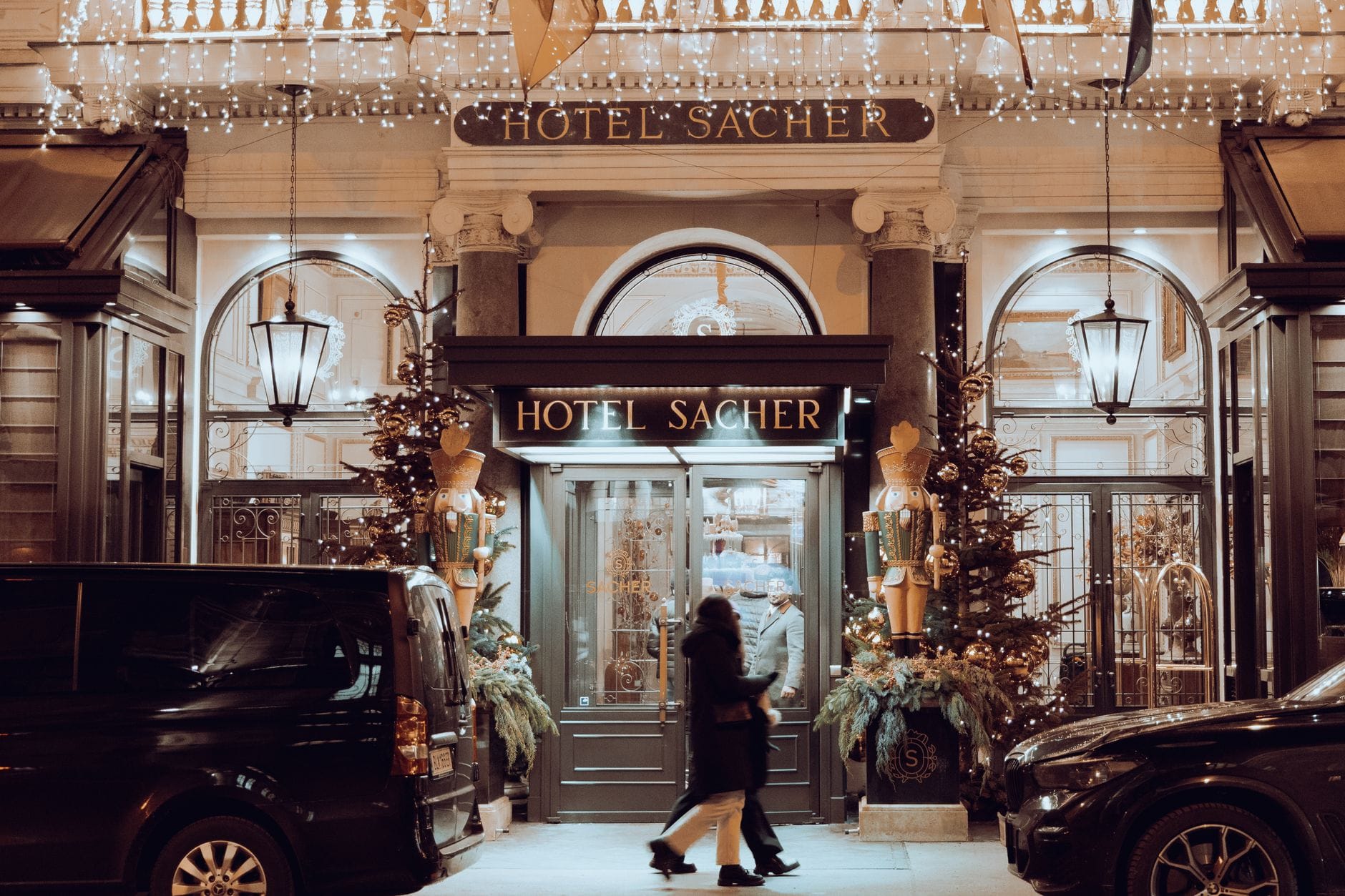 Nächtlicher Blick auf den Eingang des Hotel Sacher in Wien, mit beleuchtetem Schriftzug und dekorativen Lichtern, die Besucher willkommen heißen.