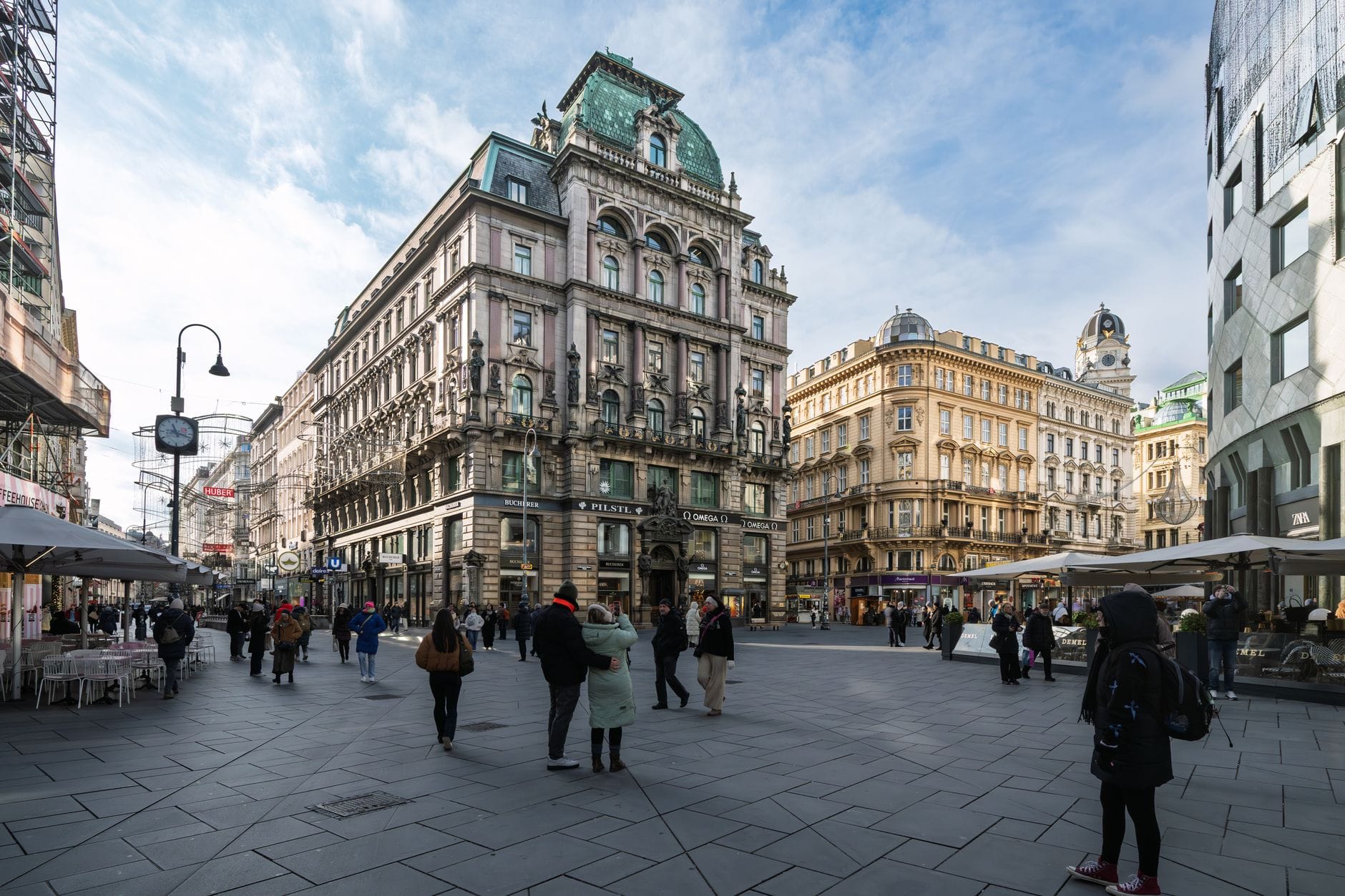 Eine belebte Einkaufsstraße im Wiener Stadtzentrum mit eleganten historischen Gebäuden, Einkäufern und Cafés entlang der Fußgängerzone an einem klaren Tag.