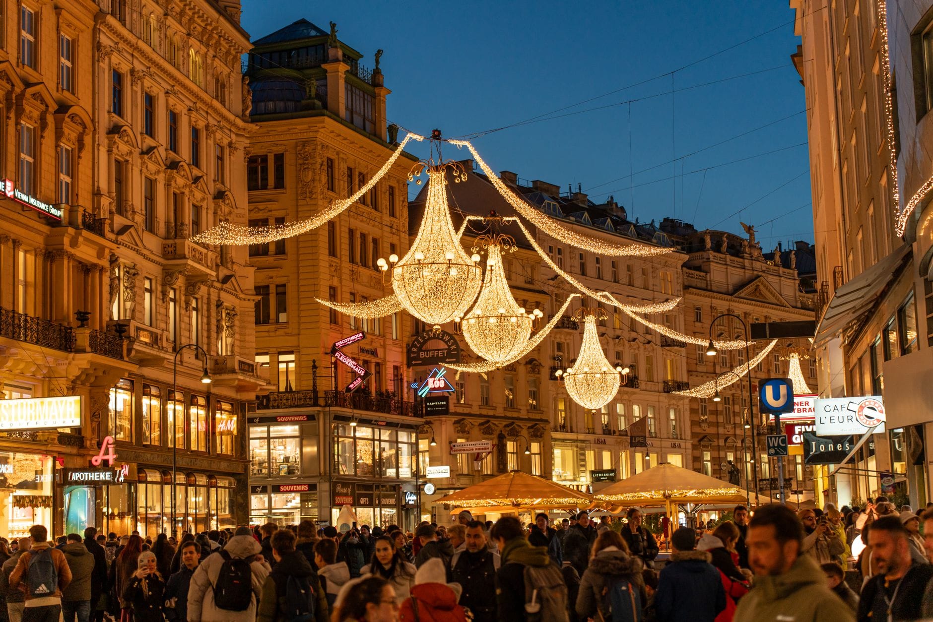 Belebte Wiener Einkaufsstraße in der Abenddämmerung mit prunkvollen Kronleuchtern, historischen Fassaden und warmbeleuchteten Schaufenstern – festlich und einladend.