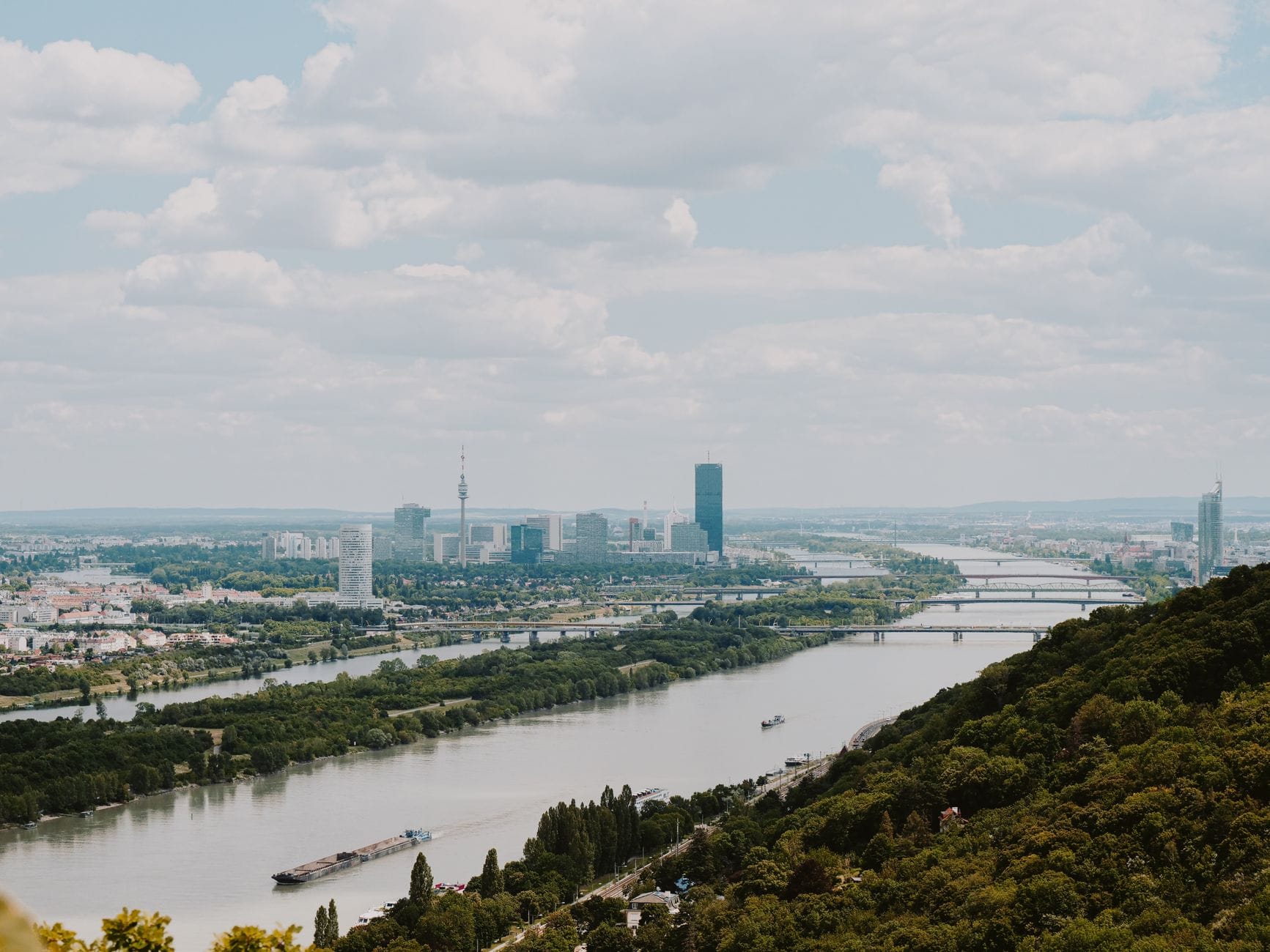 Panoramablick bei Tag über die Donau in Wien mit moderner Skyline, mehreren Brücken und grünen Hügeln im Vordergrund.