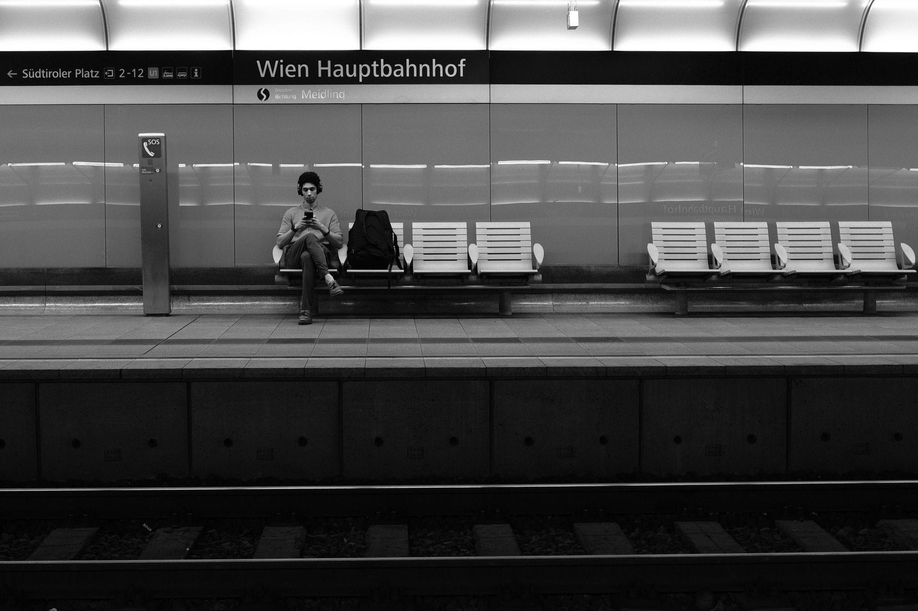 Monochromes Bild eines nahezu leeren Bahnsteigs im Wien Hauptbahnhof, mit einem sitzenden Fahrgast und gut sichtbarer Stationsbeschilderung.