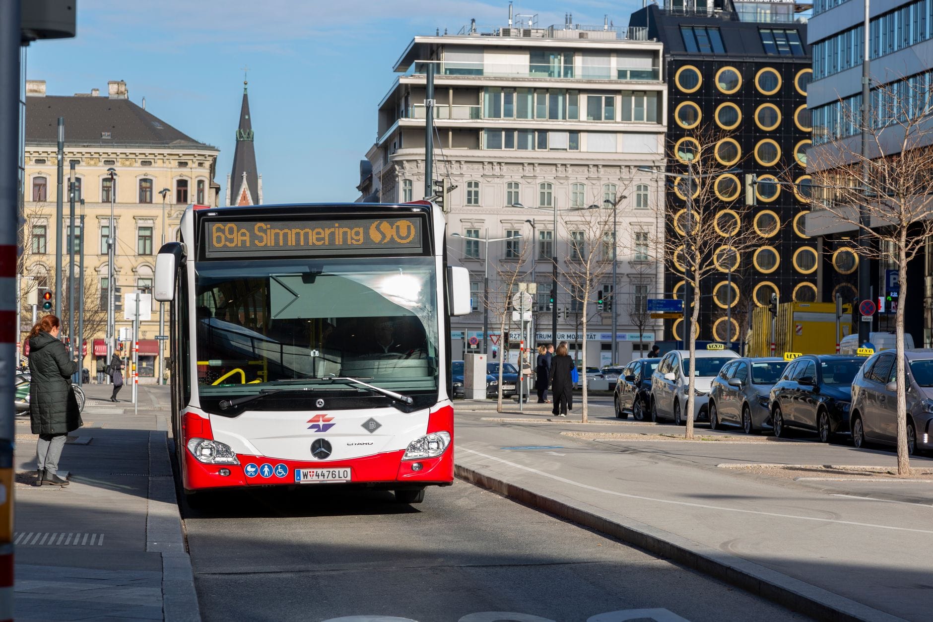 Straße in Wien mit einem rot-weißen Linienbus und modernen Gebäuden im Hintergrund an einem sonnigen Tag
