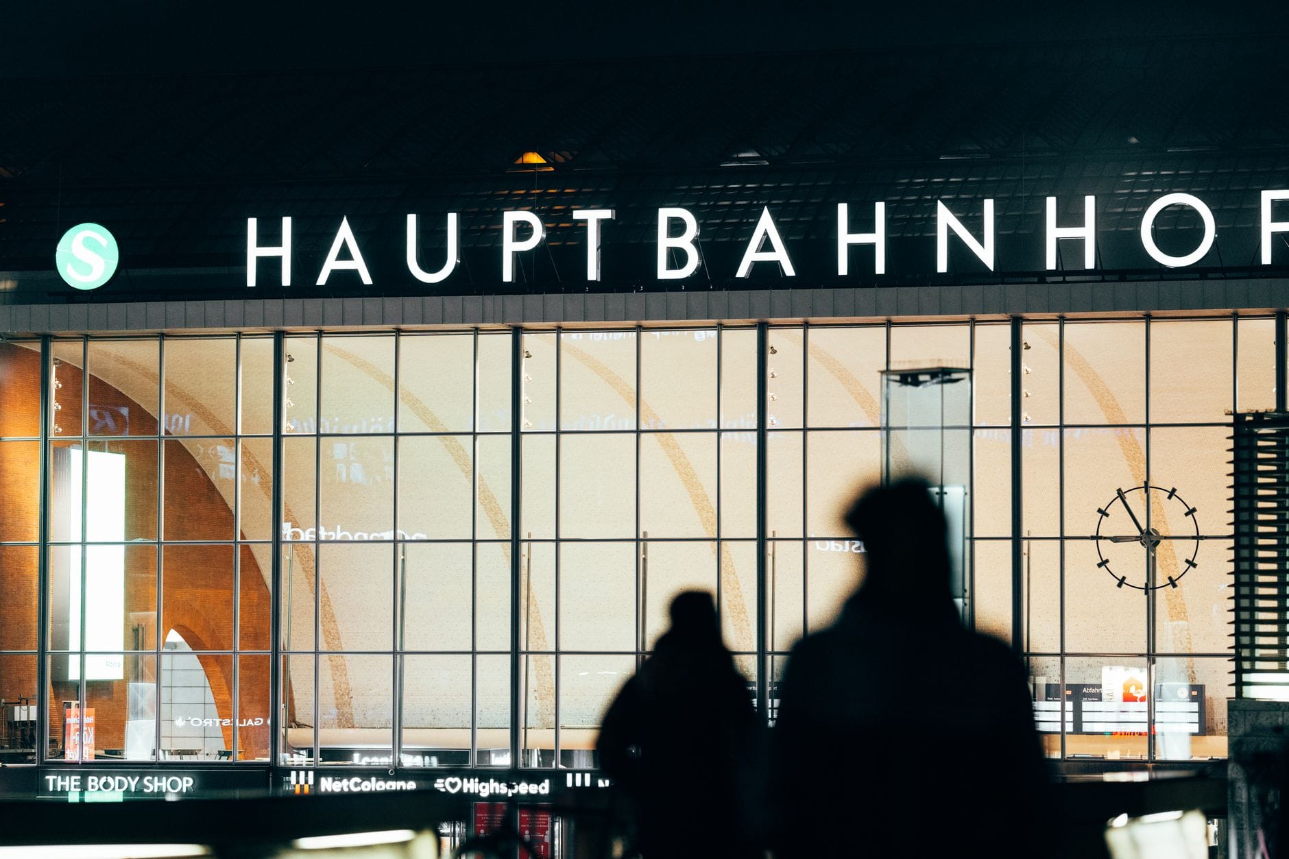 Vista nocturna de la entrada de la estación Hauptbahnhof con figuras en silueta y gran fachada de cristal.