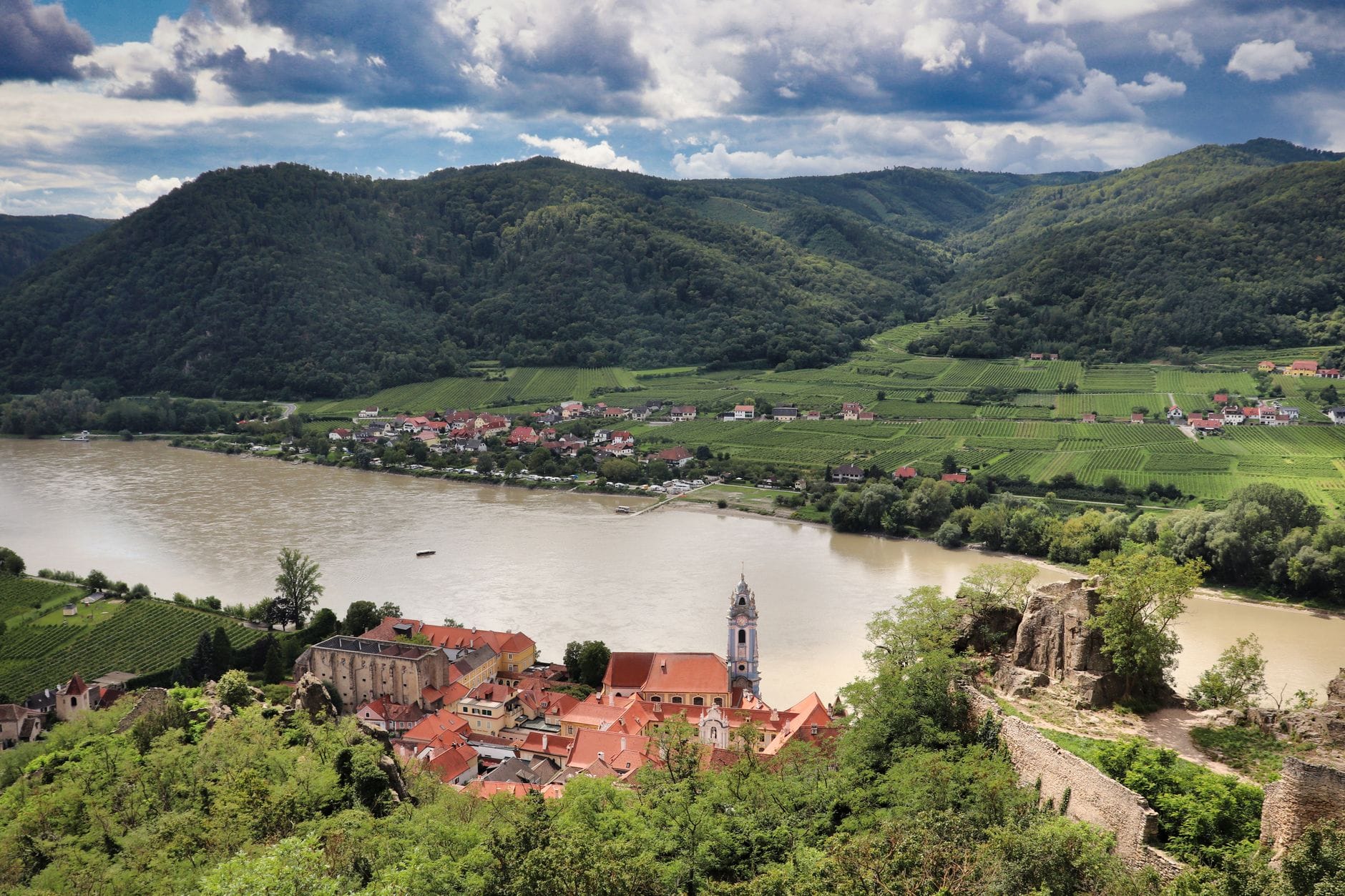 Panoramablick über das Wachautal mit dem blauen Kirchturm von Dürnstein, der Donau, terrassierten Weinhängen und der Burgruine vor dramatischem Himmel.
