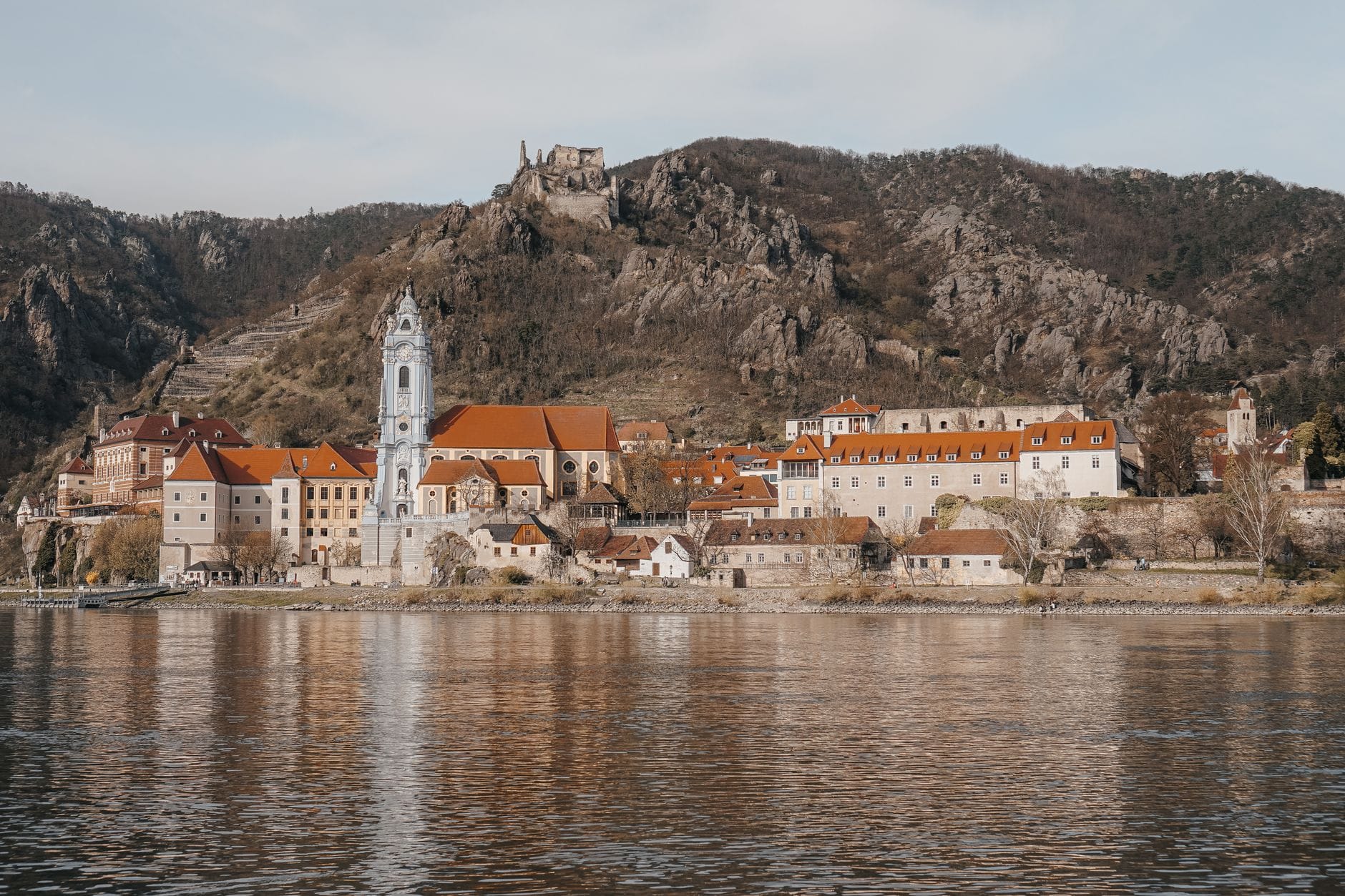 Vista de Dürnstein con su torre de iglesia azul, pintorescos edificios del pueblo, viñedos y ruinas del castillo en lo alto de la colina, vistas desde el río Danubio bajo cielos despejados.